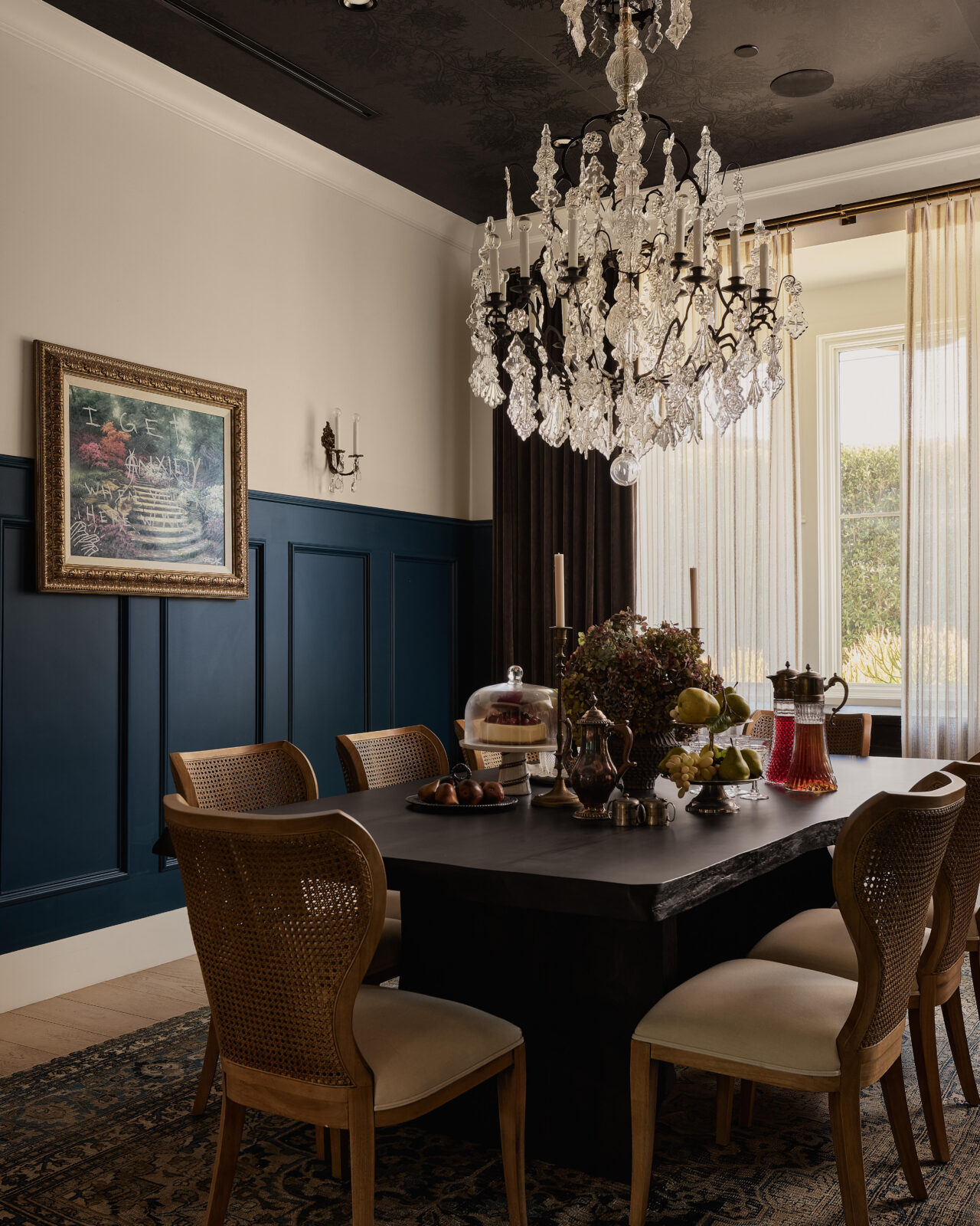 Formal dining room wrapped in deep blue paneled walls and a dark ceiling, setting the stage for the oversized vintage Parisian chandelier that hangs over the dining table and cane-back chairs.
