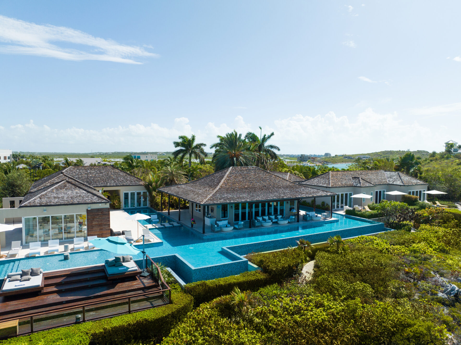 The expansive infinity-edge pool stretches between multiple pavilions, with built-in ledges, deep-blue tile, and surrounding open-air sun loungers that connect the water to the house.