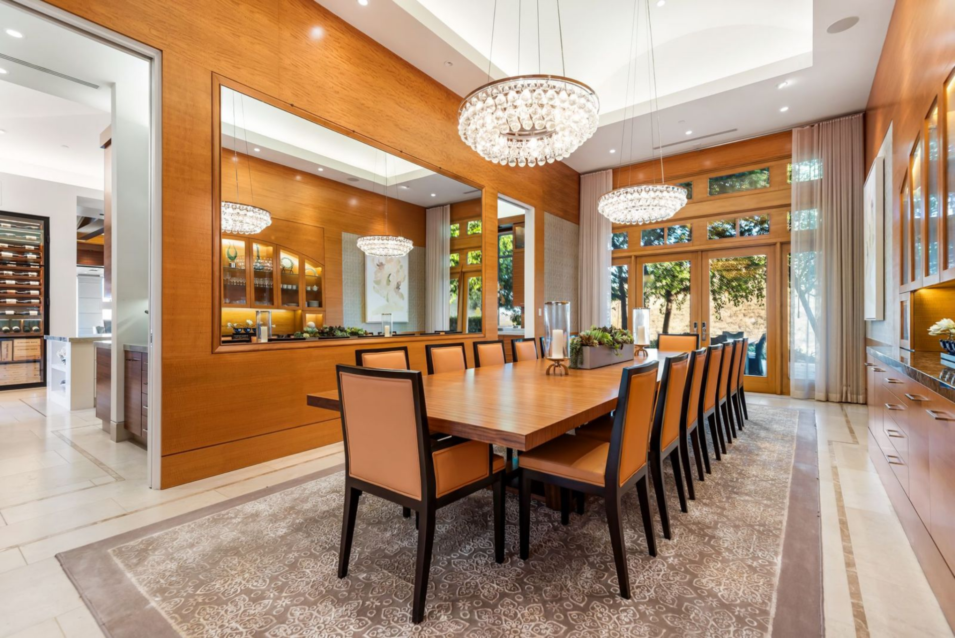 Warm wood paneling wraps the dining room where a long dining table is positioned beneath two crystal chandeliers.