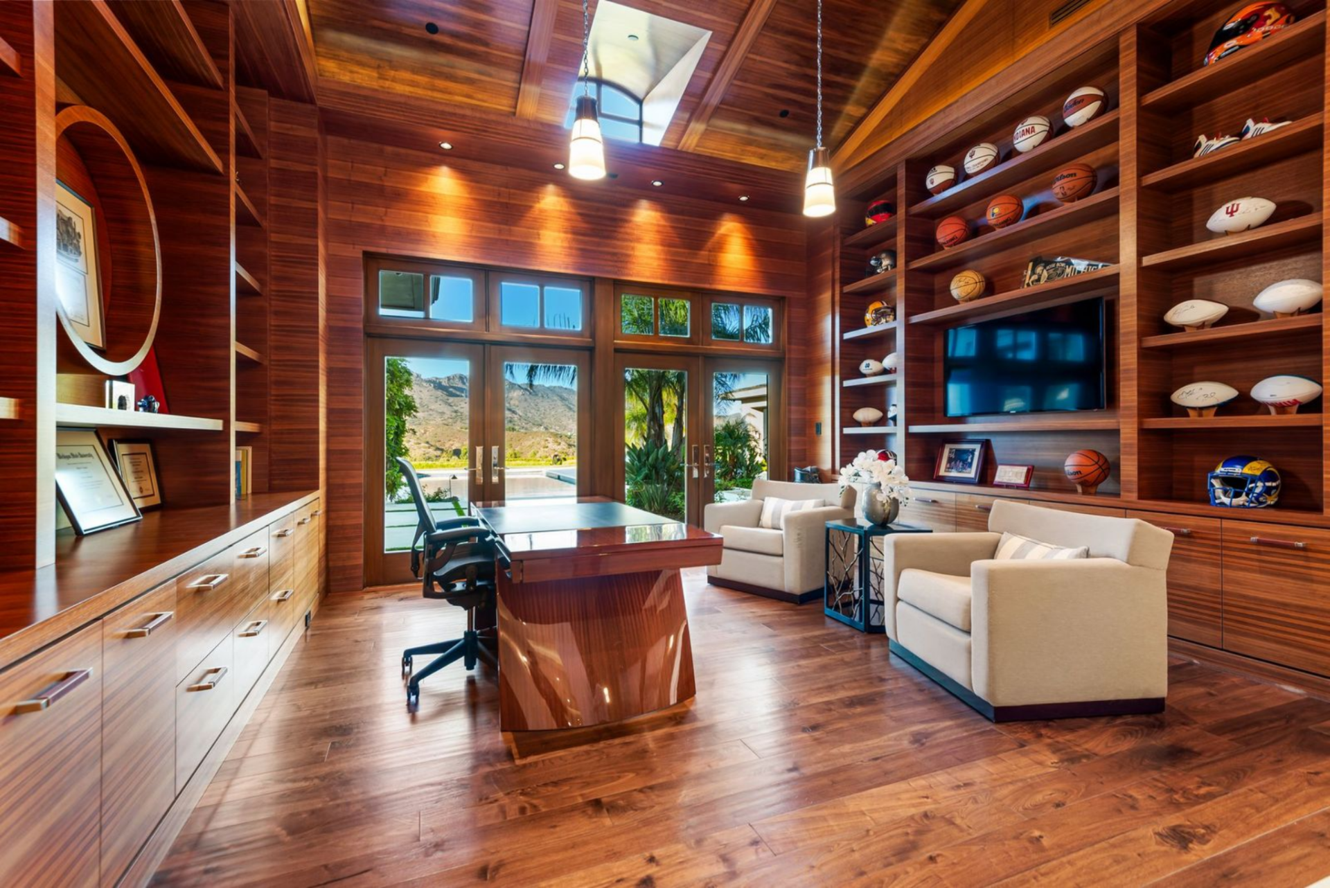 A sculptural desk sits across from dark wood shelving displaying signed footballs, basketballs, and sports helmets.