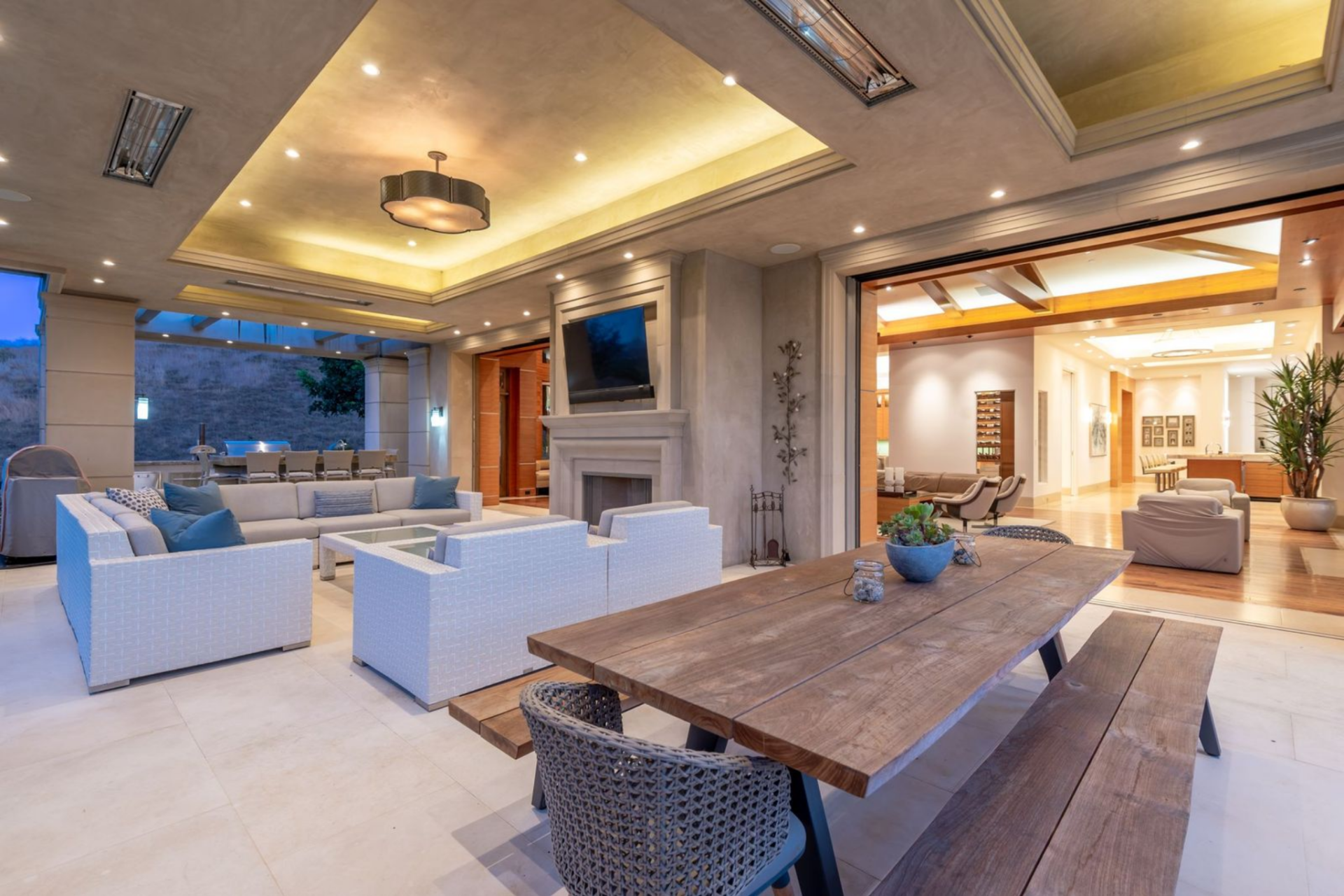 Three sections sit in an outdoor patio: an outdoor dining table, a seating area centered around a fireplace with a mounted television above it, and an outdoor kitchen