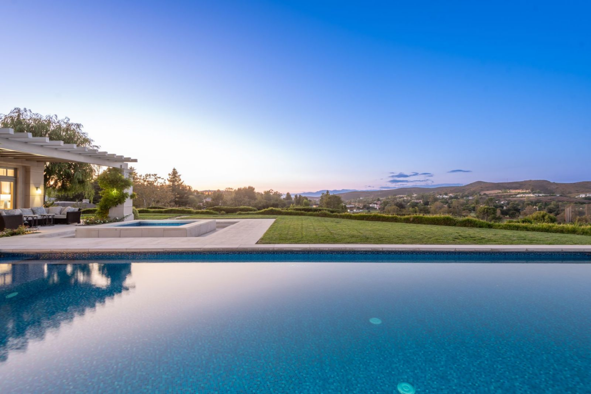 A broad lawn and clean stone hardscaping surround a pool and spa looking out to the horizon at sunset.