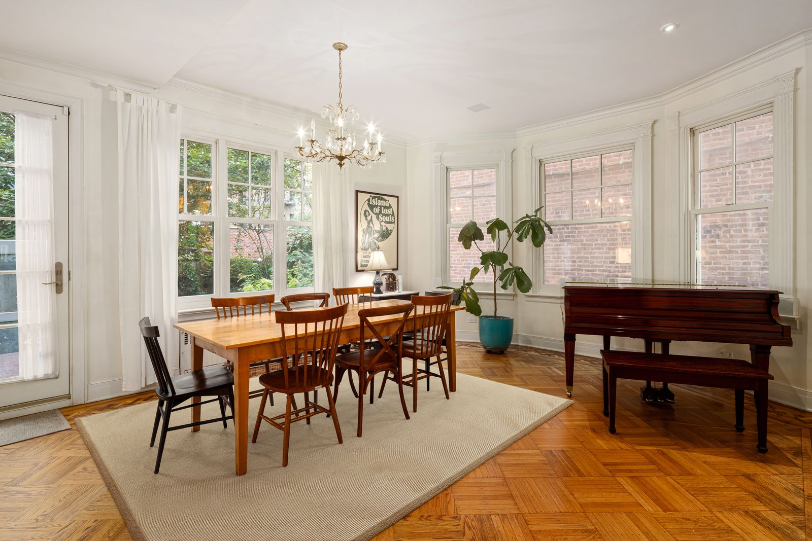 Dining room with a wood dining table centered beneath a traditional chandelier, with mismatched chairs and a piano in the corner.