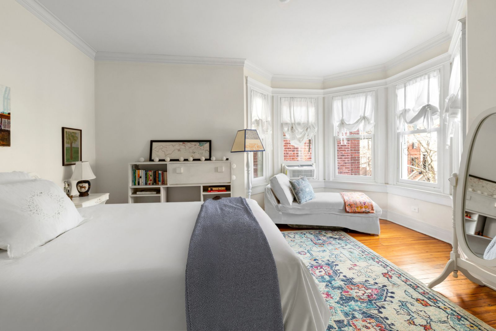 Bedroom with a bed on top of a vivid rug, books lining a bookshelf along one wall, and a corner window nook with a lounge chair.