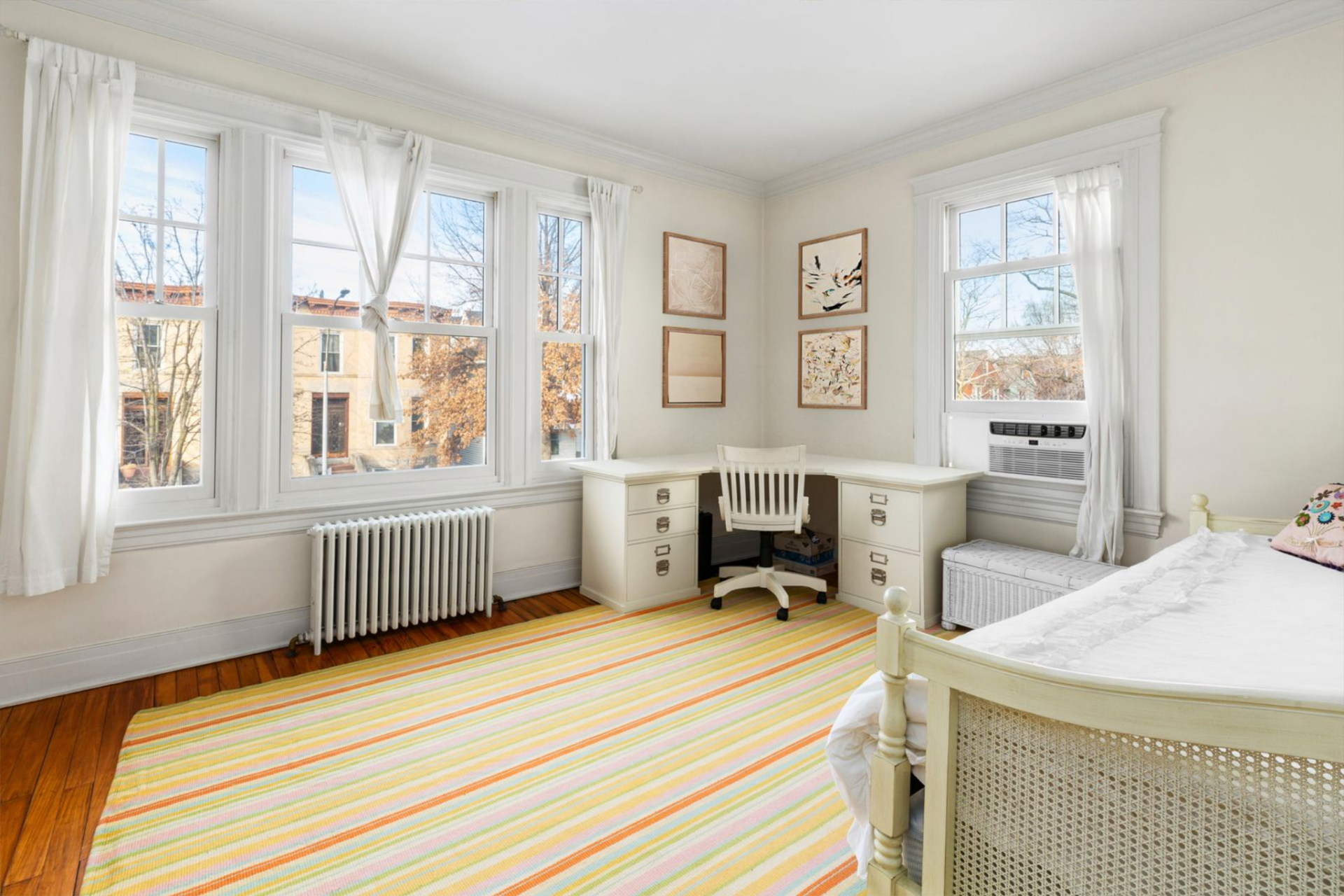 Bedroom with a corner desk positioned between the room's windows on top of a bright stripped rug.