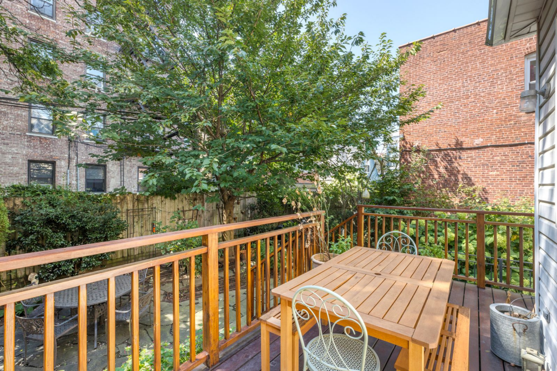 A raised wooden deck extends directly off the rear of the house, set up for outdoor meals with a simple table and chairs framed by wood railings and leafy views of the yard. 
