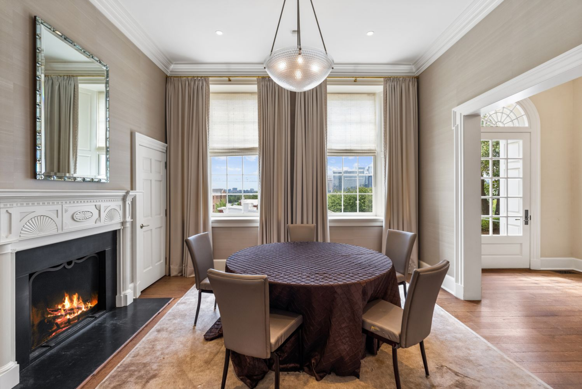 Room with a carved white fireplace with classical detailing and tall windows dressed in full-length drapery with a round table in the middle.