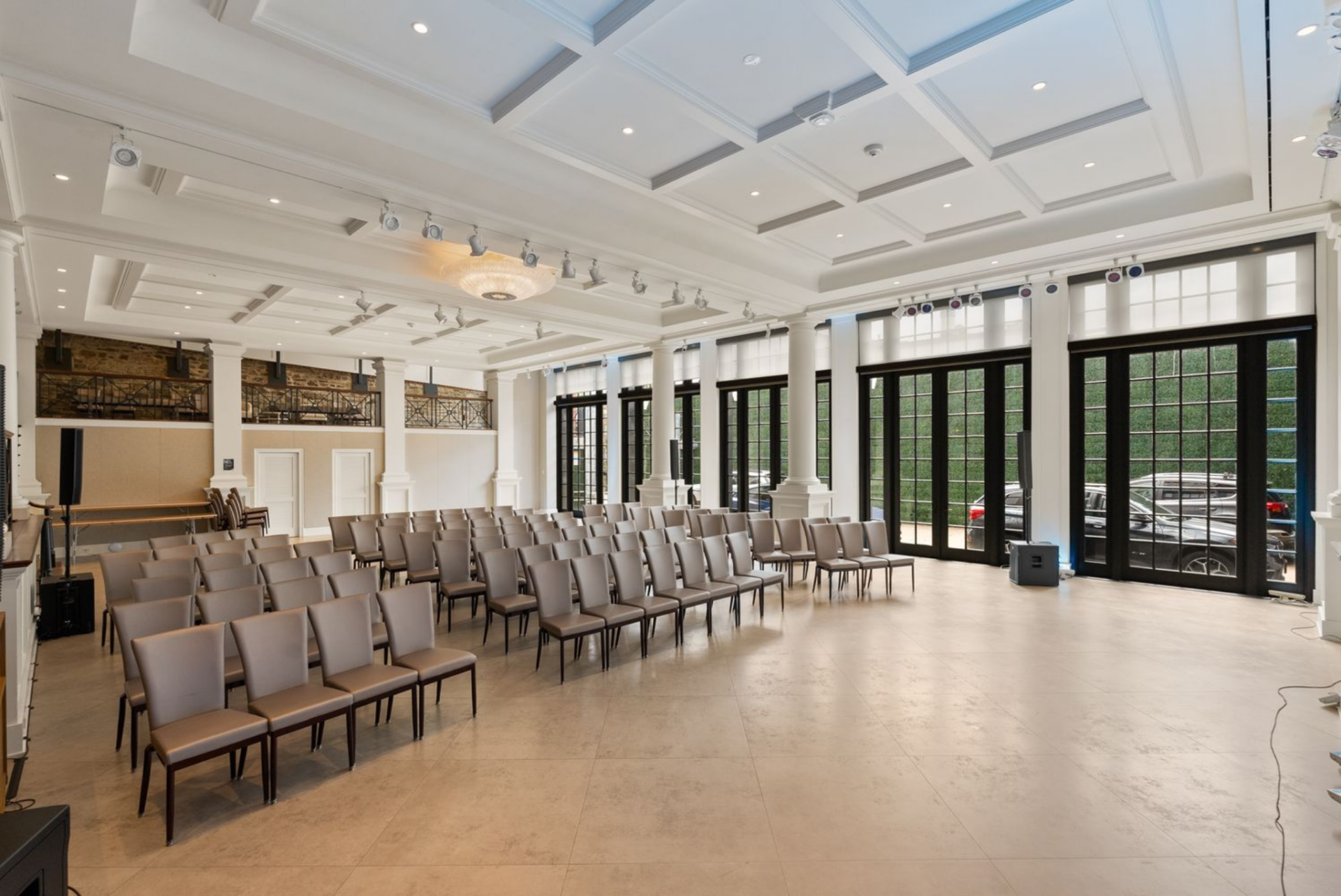 A grand assembly with rows of seating arranged beneath a coffered ceiling, punctuated by classical columns, discreet track lighting, and a central chandelier.