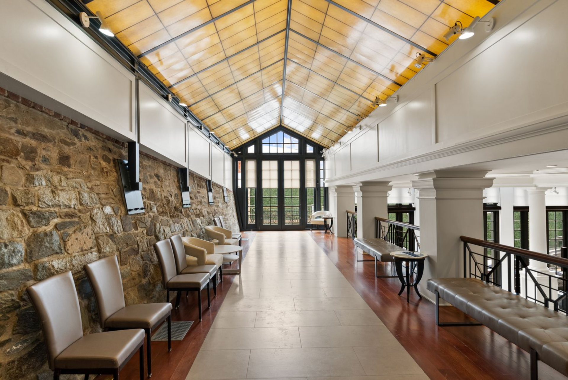 A balcony overlooking a grand hall, lined with exposed stone walls on one side and white classical detailing on the other. 