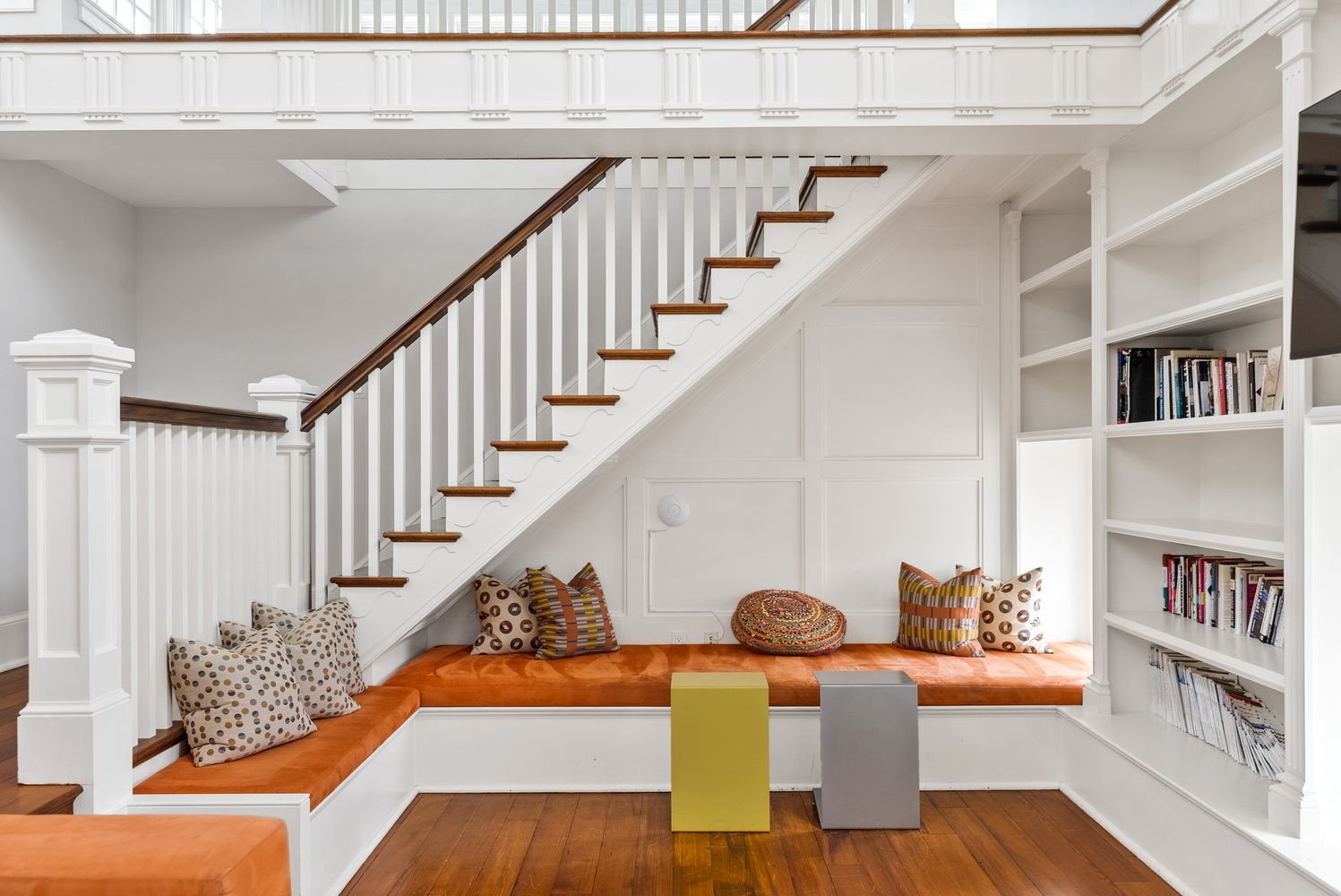 Library seating nook beneath staircase with orange cushions.