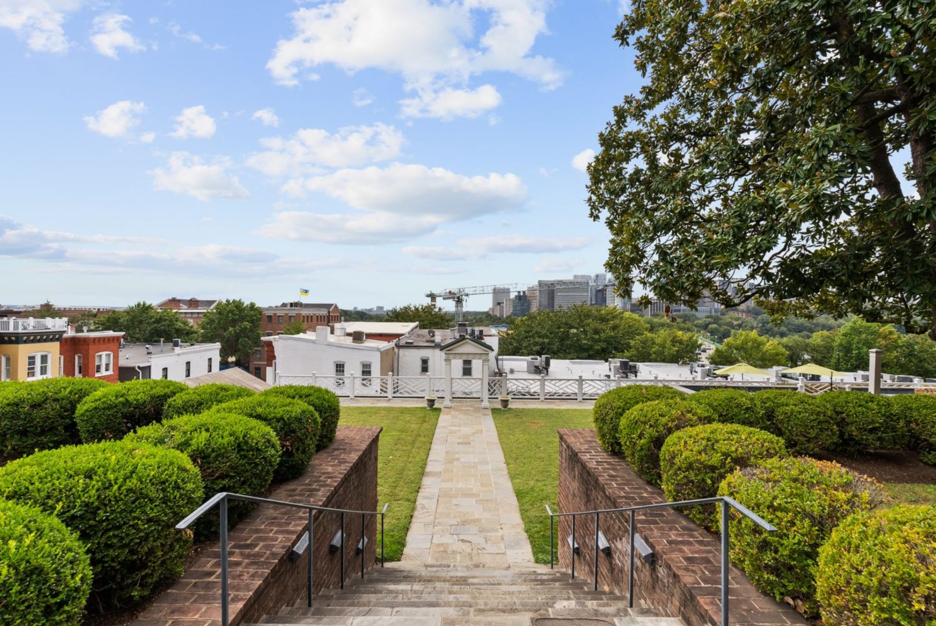 A central stone path leads away from the house, framed by trimmed hedges and brick walls overlooking the rooftops.