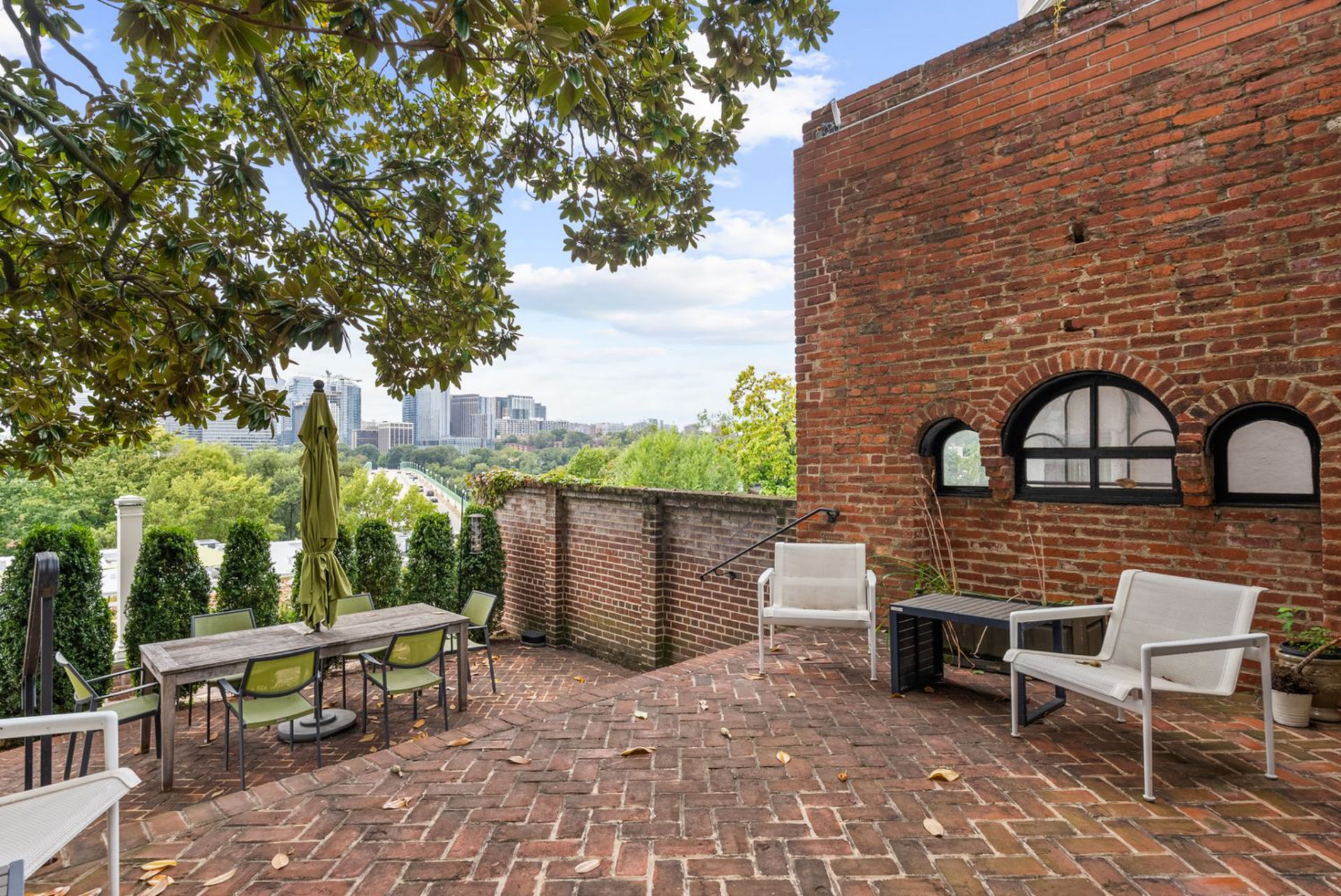 A brick-paved terrace arranged for outdoor dining, with a long table positioned beneath mature trees next to the home’s historic brick façade. 