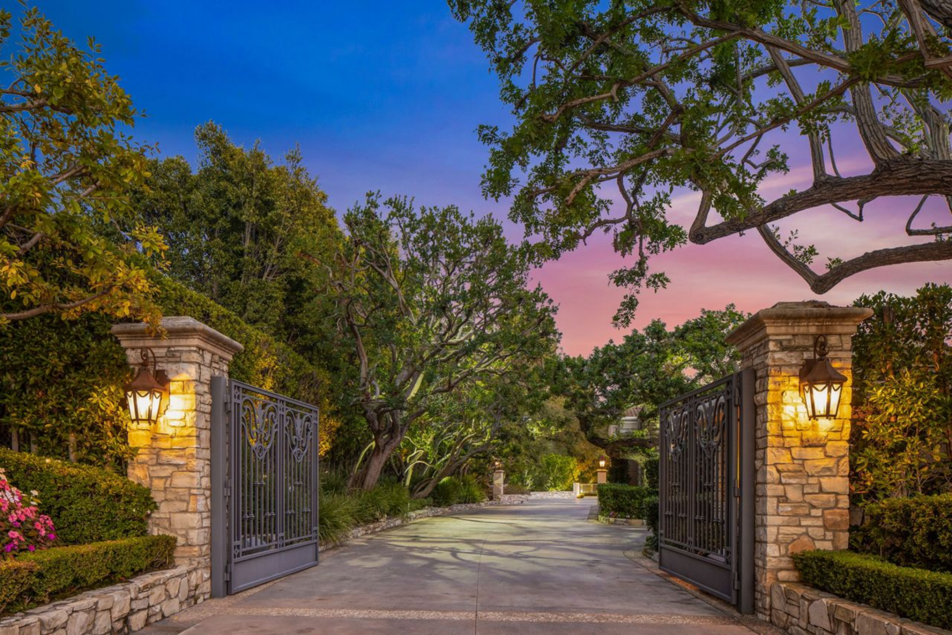 Johnny Carson’s former Bel Air mansion begins at a gated entry marked by stone pillars and wrought-iron doors, opening to a road lined with trees. 