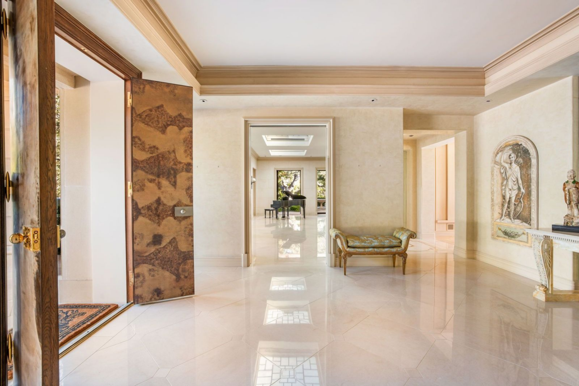 A grand foyer opens across polished stone floors and layered crown moldings guide your eye toward a sunlit room at the end of the hall with a grand piano.
