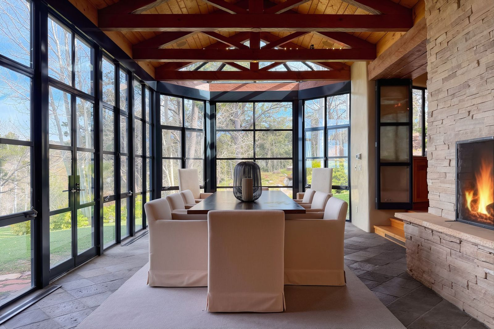 Dining space wrapped almost entirely in glass with a long rectangular dining table set low surrounded by upholstered chairs next to a stone fireplace.
