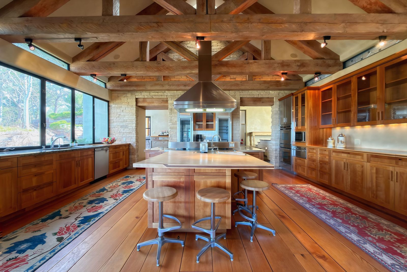 The kitchen has wood cabinetry that runs wall to wall, with open shelving and glass-front uppers that frame a stainless steel range hood dropping down from the ceiling over a thick butcher-block island.