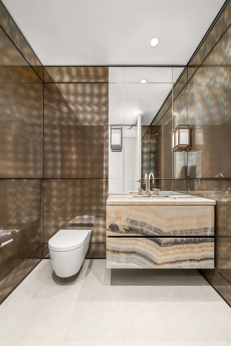 The powder room makes a strong first impression with a sculptural vanity wrapped in richly toned stone, metallic wall panels, and soft, directional lighting that add depth without overwhelming the compact space.