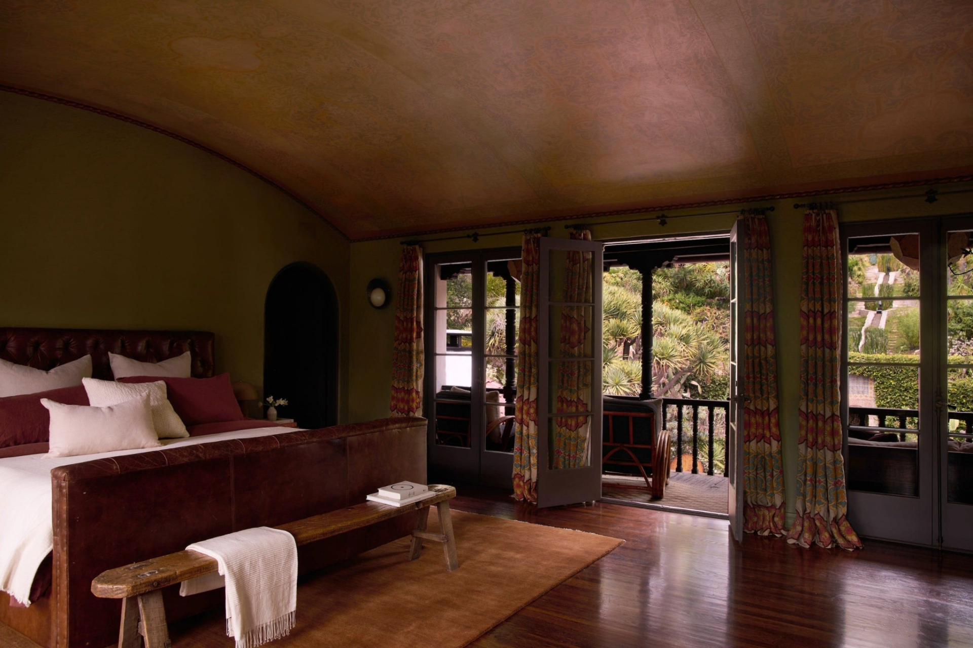 The bedroom features a vaulted, arched ceiling finished in a hand-applied plaster, while French doors framed by patterned drapery open to the balcony and bring the hillside greenery into the room. 