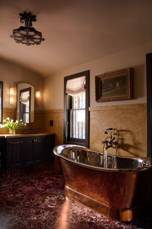 In the bath, richly veined stone floors, a freestanding metal soaking tub, and custom cabinetry are paired with classic sconces and arched mirrors.