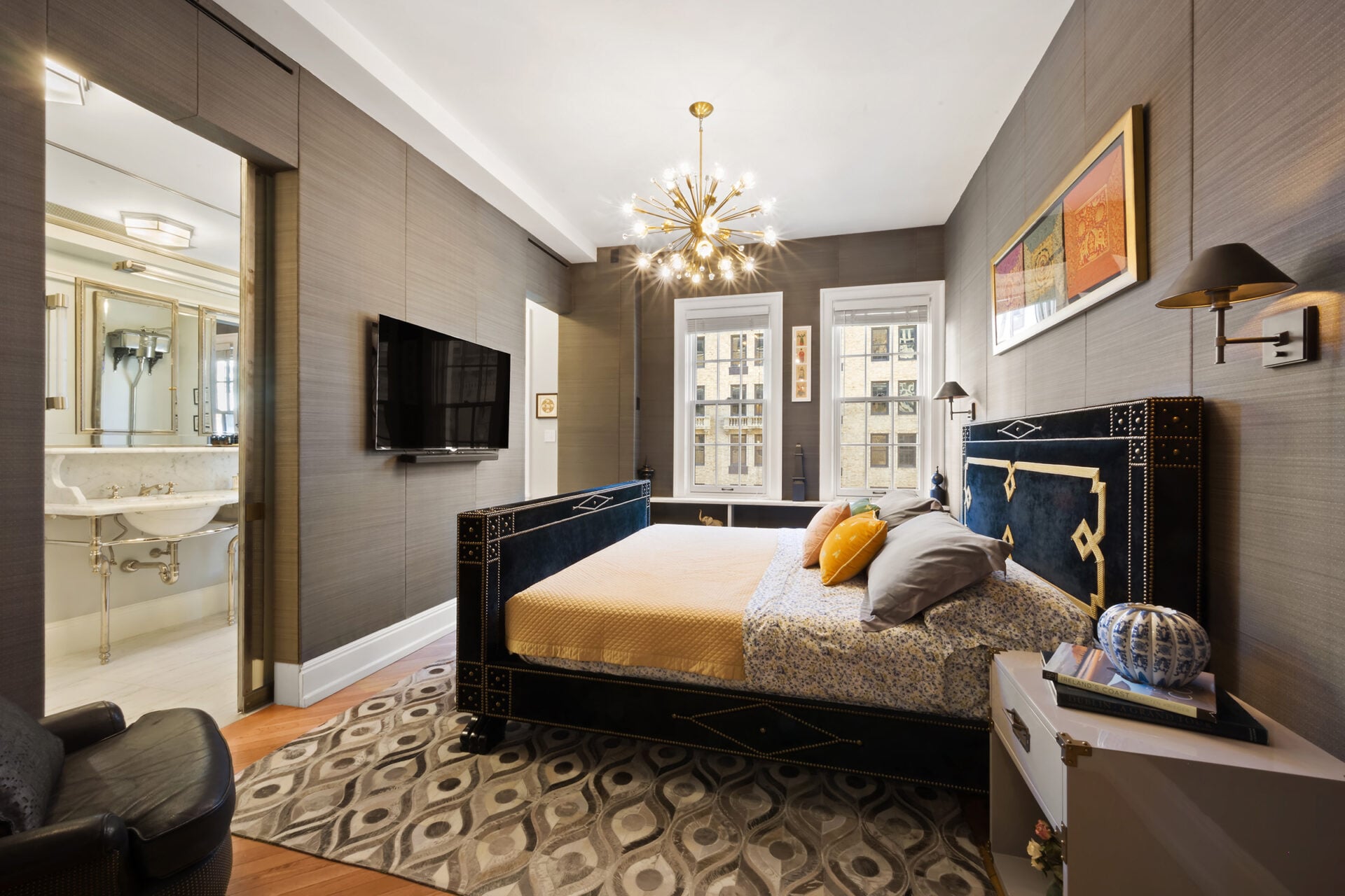 Up on the second floor, the primary suite's bedroom opens with tall ceilings, large windows, and a large bed sitting in the center of the room facing a mounted TV.