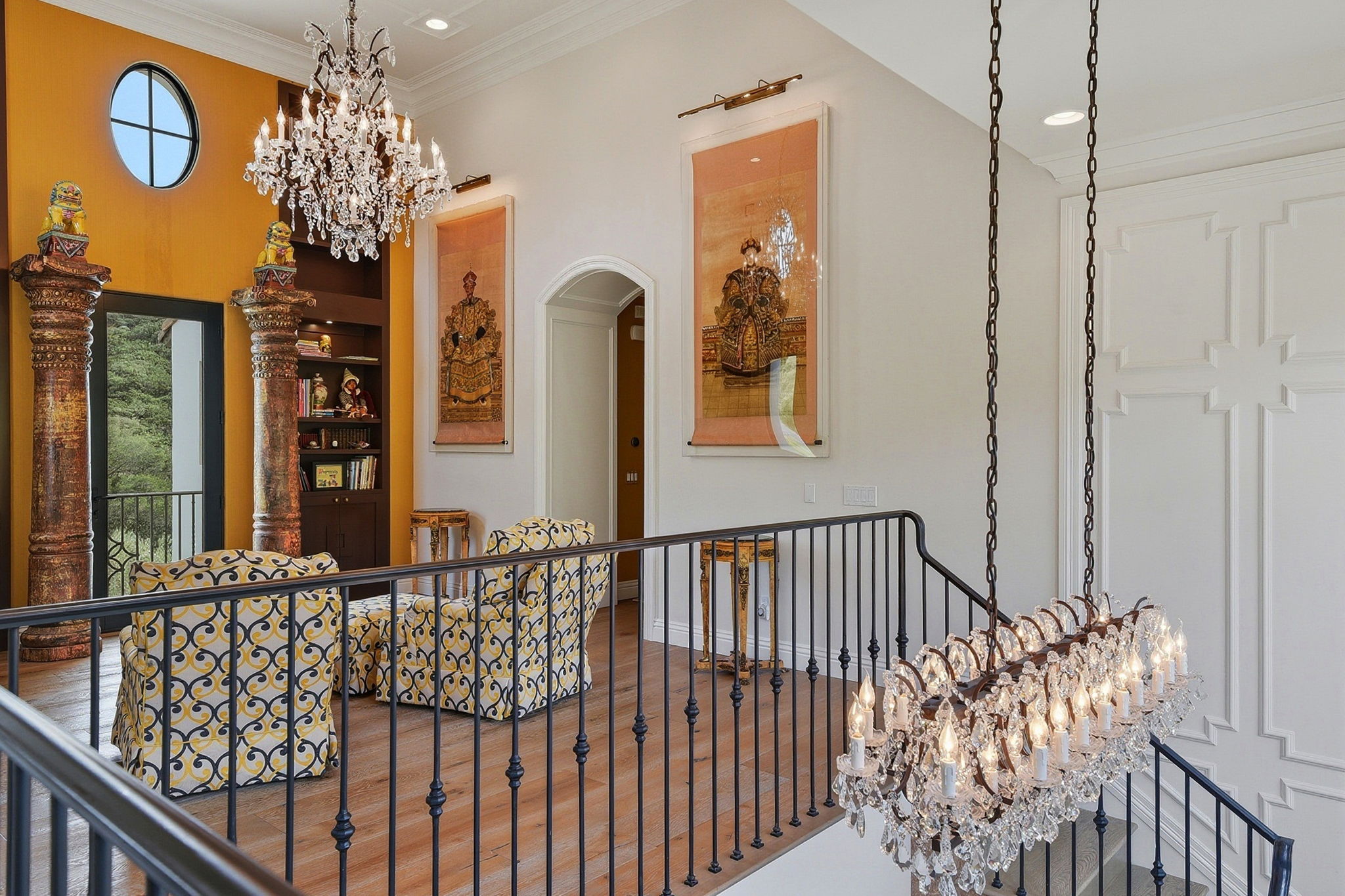 Framed by wrought-iron railings and illuminated by a cascading crystal chandelier, the space includes two patterned armchairs that sit facing a bold saffron accent wall, framed by columns and large-scale art panels that add depth and personality.