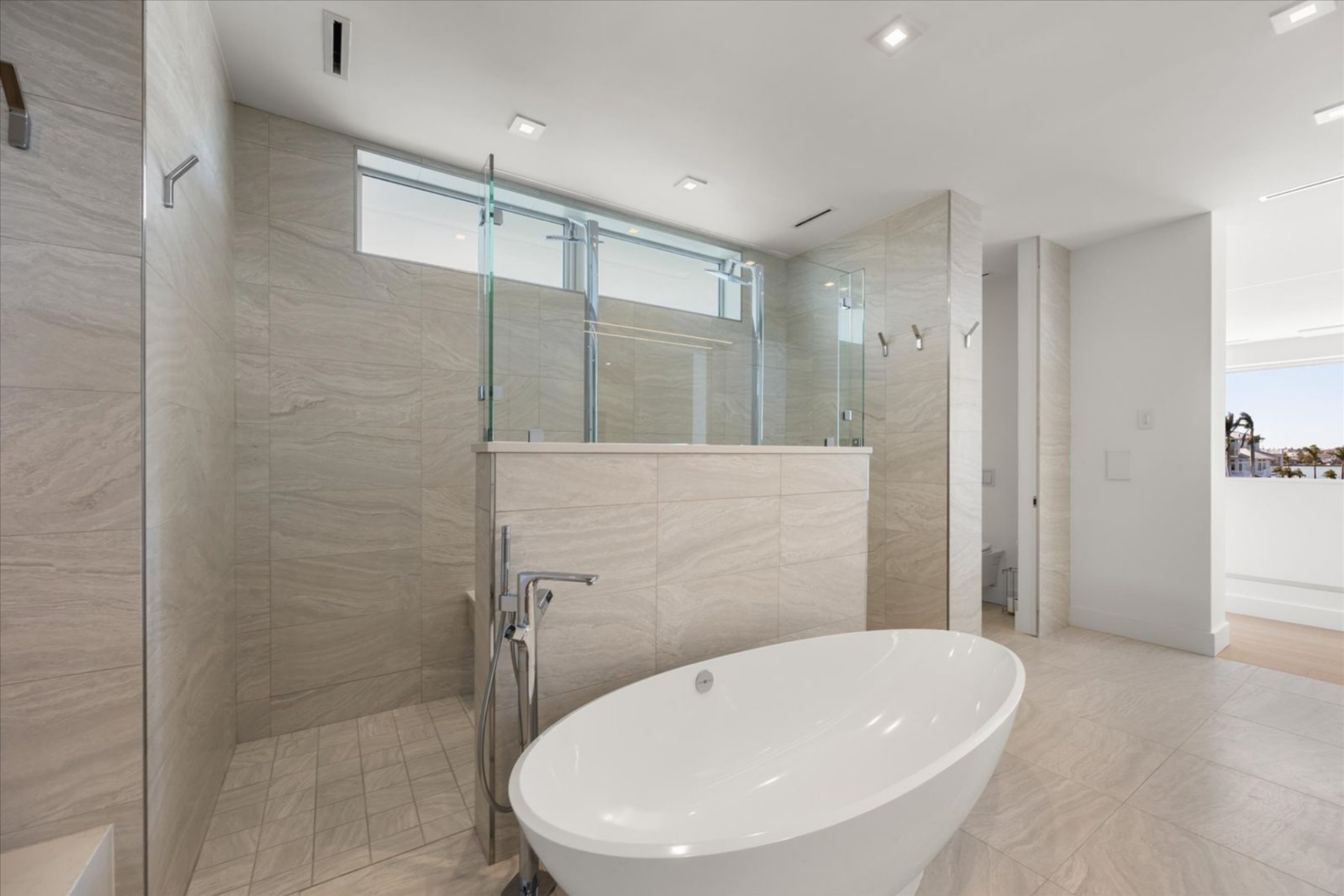 Moving deeper into the suite, the layout transitions into the bath where a freestanding tub is set against full-height stone walls and glass partitions that define the oversized Roman shower behind it. 