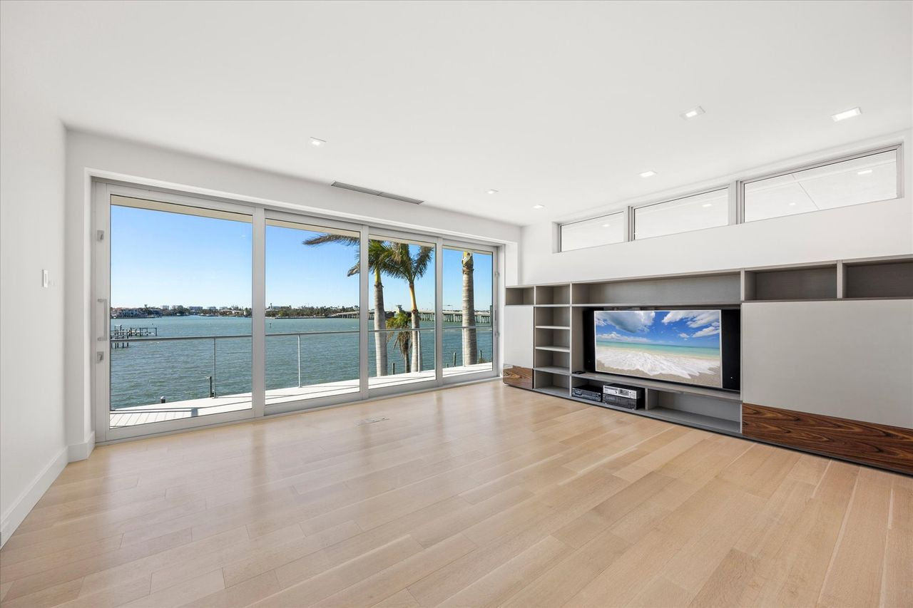 The media room is designed as a relaxed viewing space with built-in shelving and a clean, open layout, anchored by large sliding glass doors that lead directly to a terrace overlooking the water. 