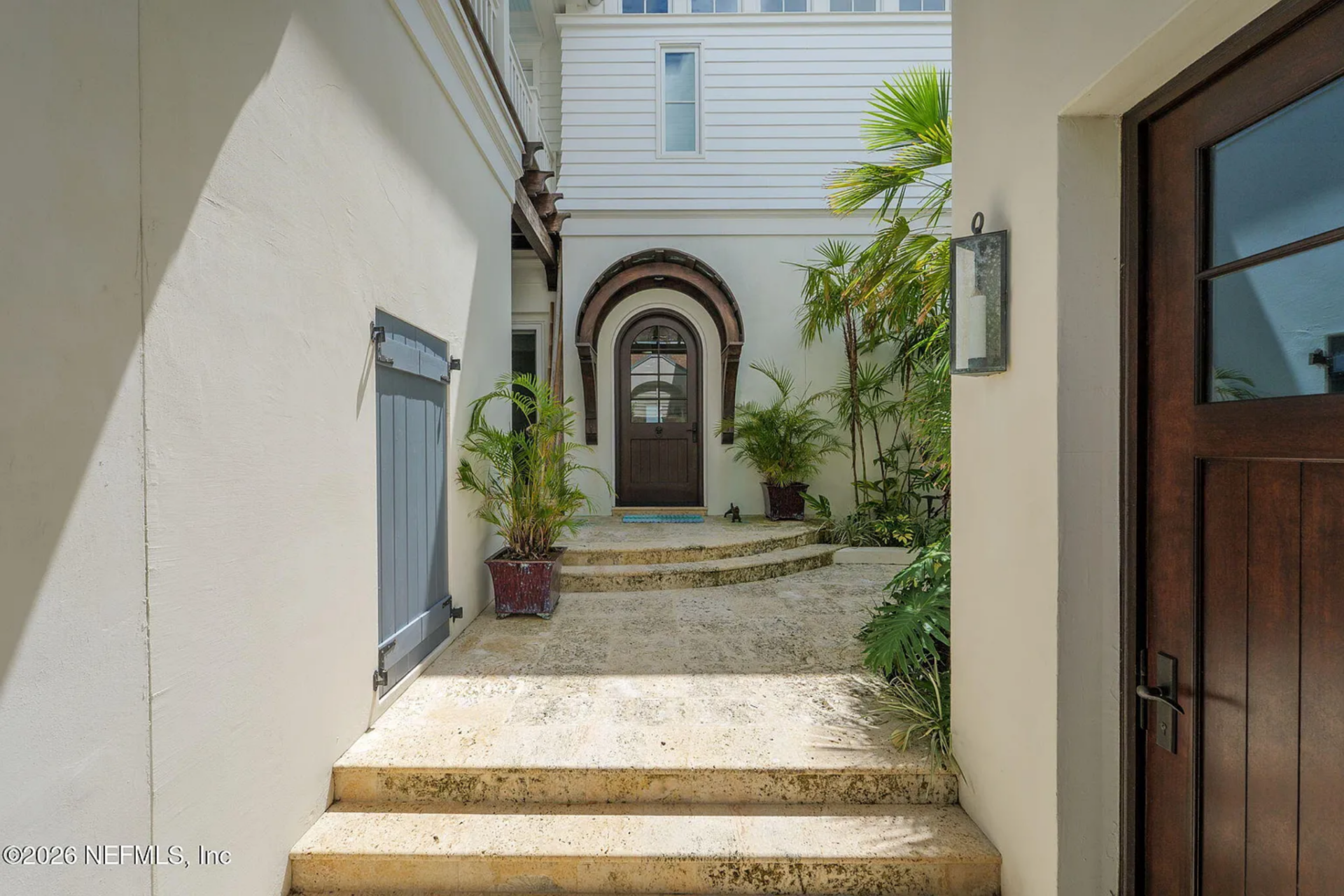 The approach to Haywood's home begins through a narrow courtyard passage lined with smooth stucco walls and tropical plants, where travertine steps lead up to an arched wooden front door framed in dark trim.