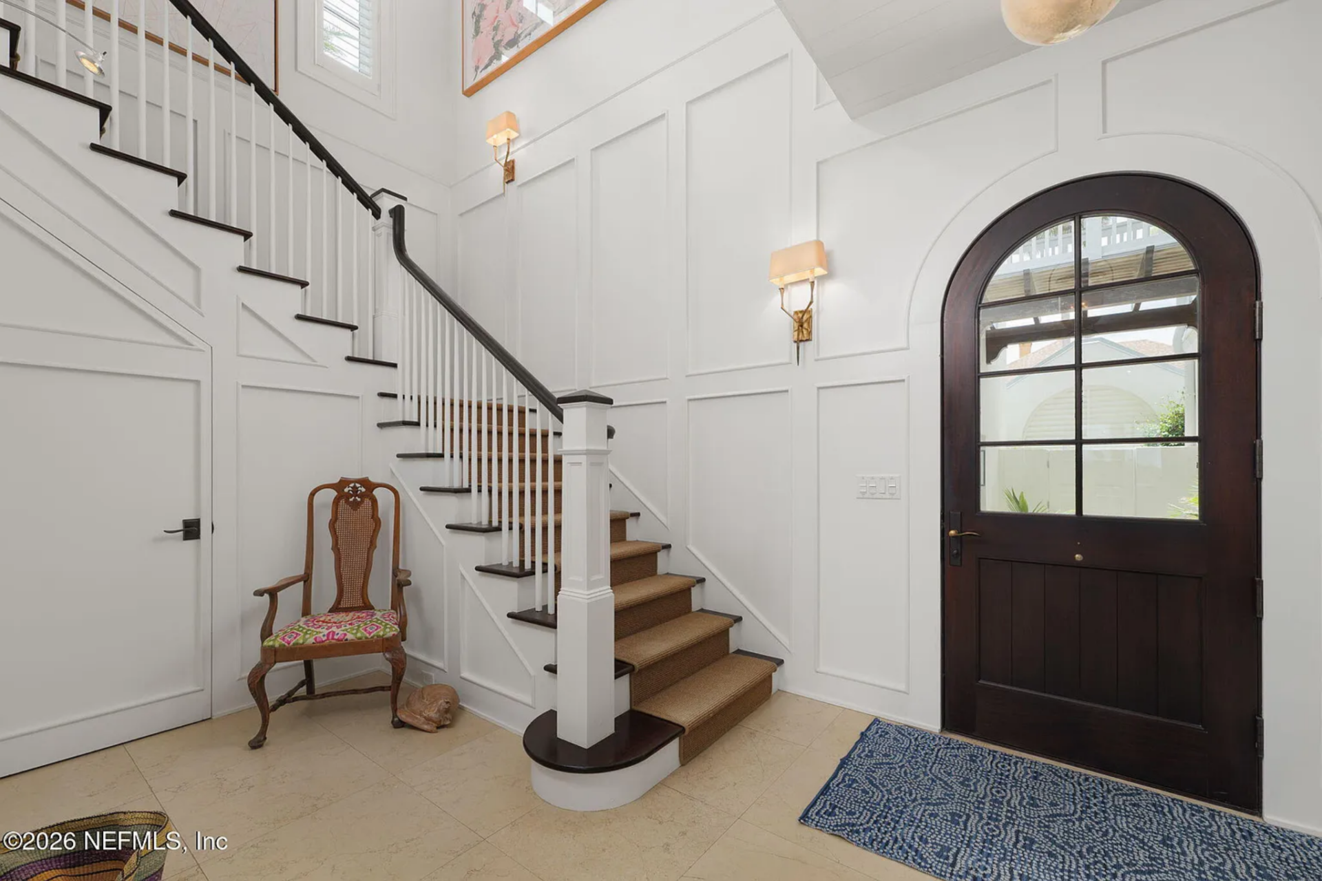 Stepping inside, the foyer opens upward with tall white paneled walls and a staircase rising along the left side, finished with dark handrails and light wood treads.