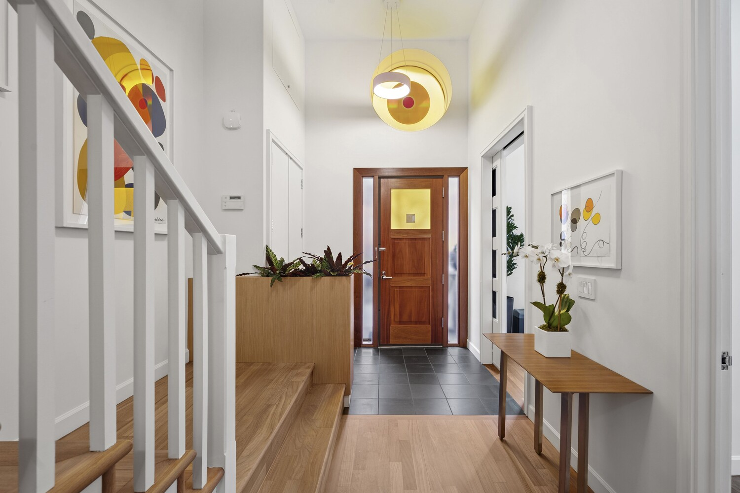A warm wood front door with a square glass inset has a sculptural yellow pendant hanging over it, while to the left, a white-painted staircase with simple vertical balusters rises cleanly along the wall.