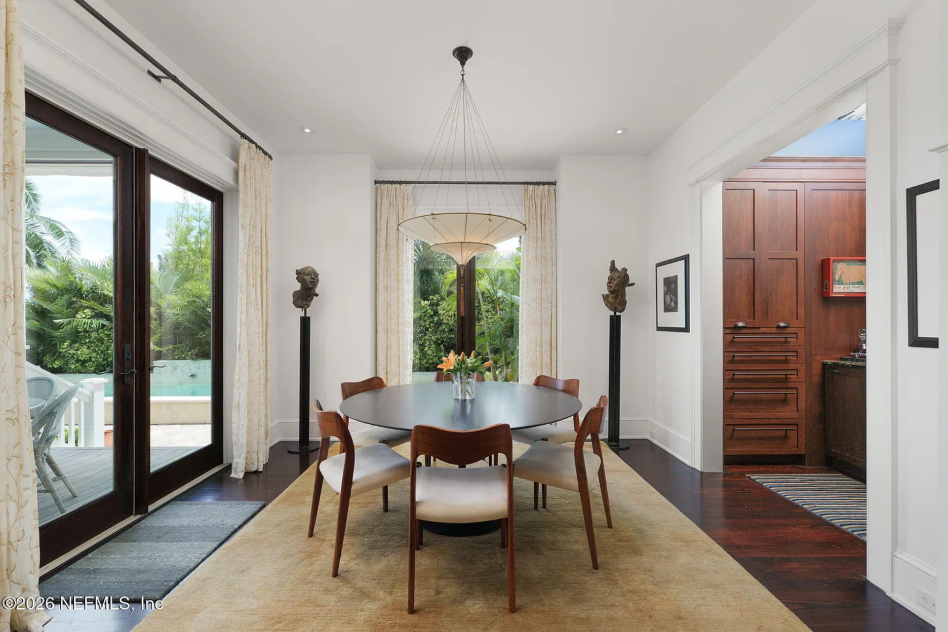 The kitchen transitions directly into the dining area, where a round dining table sits centered on a large rug beneath a suspended light fixture, framed by tall windows and glass doors that open toward greenery and the pool beyond.