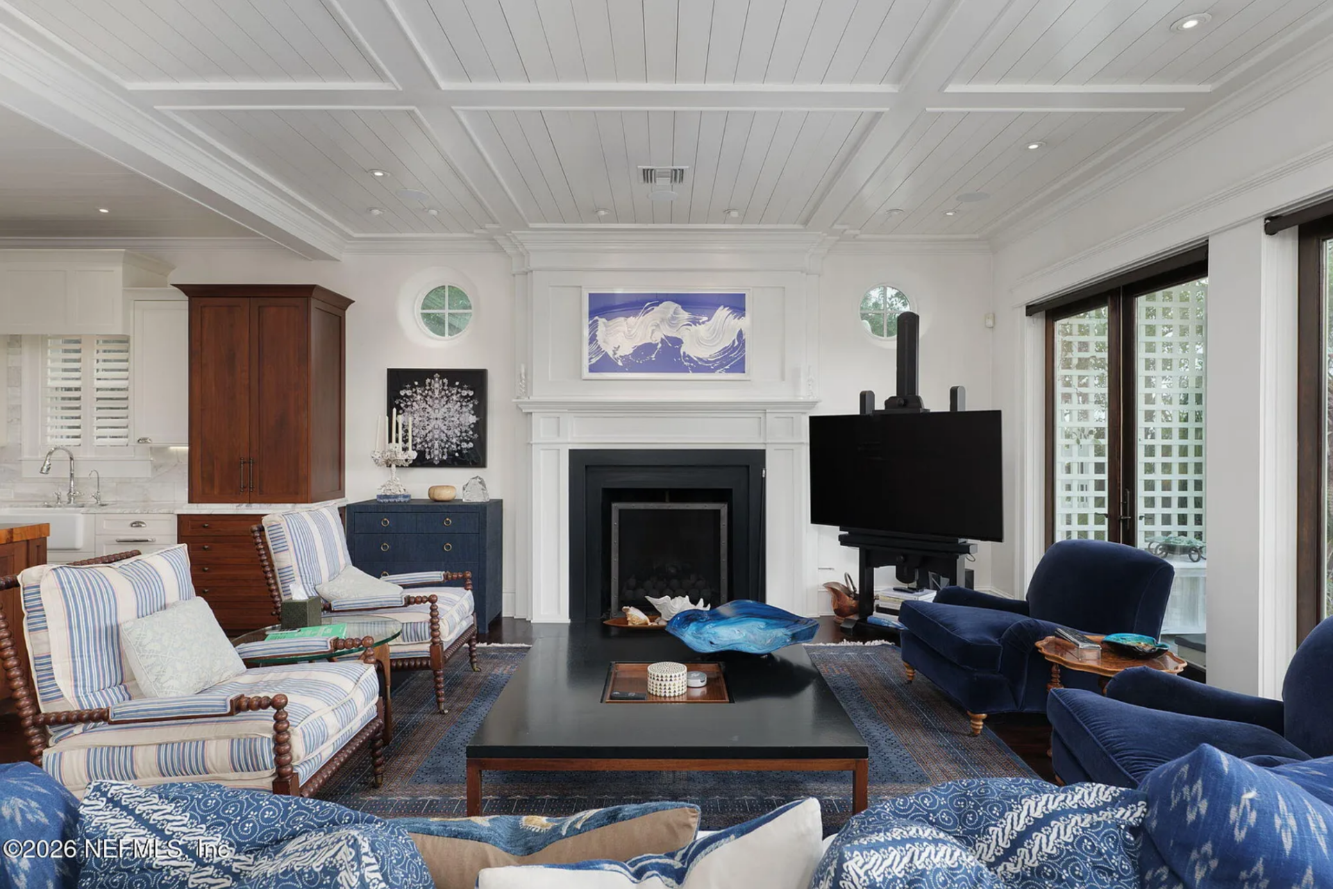 A coffered ceiling with recessed lighting stretches overhead, while a central fireplace with detailed white millwork anchors the far wall, framed by round windows that bring in filtered light.