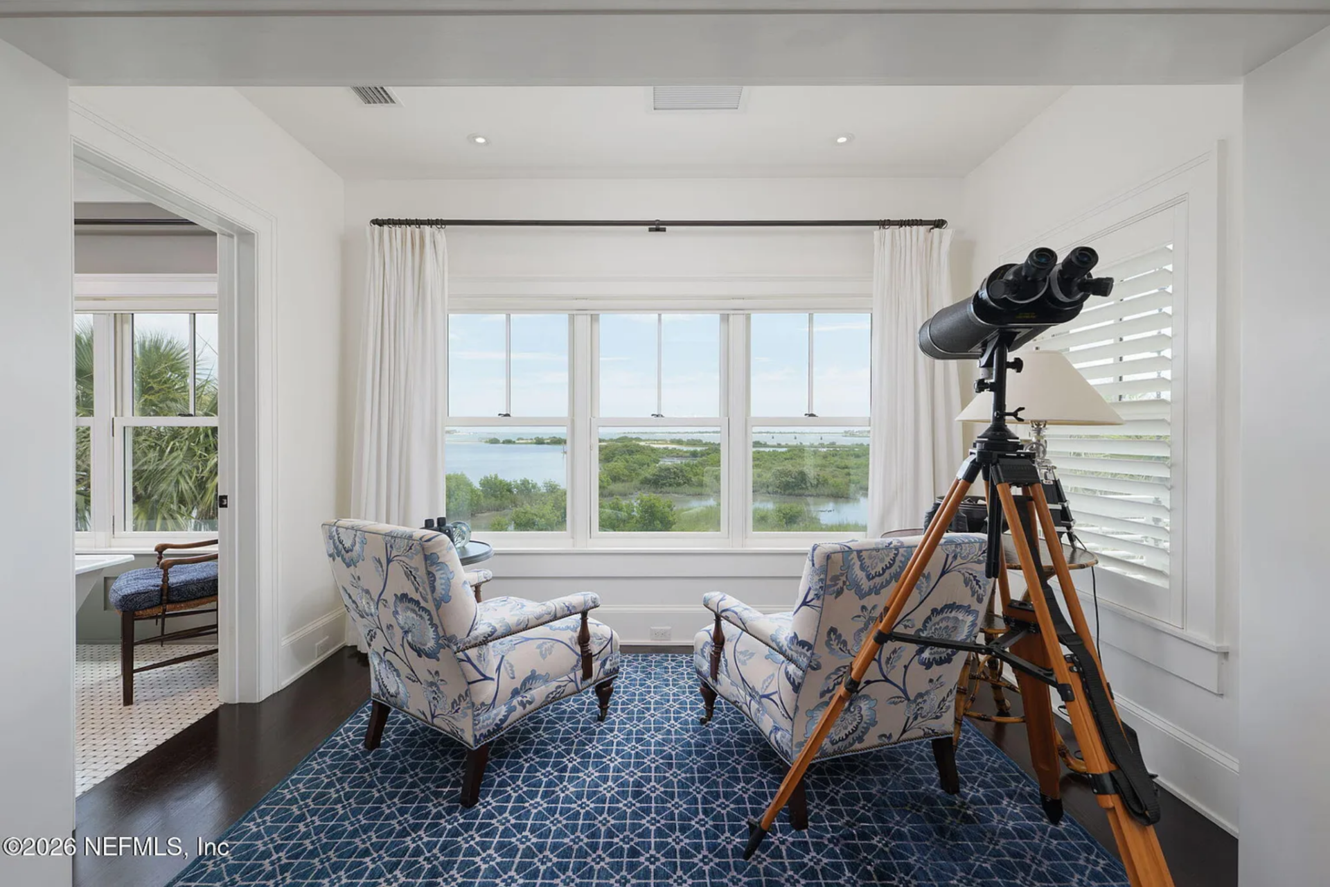 The room extends forward into a dedicated lounge area set within a window-lined alcove, where two patterned armchairs face outward toward the water.