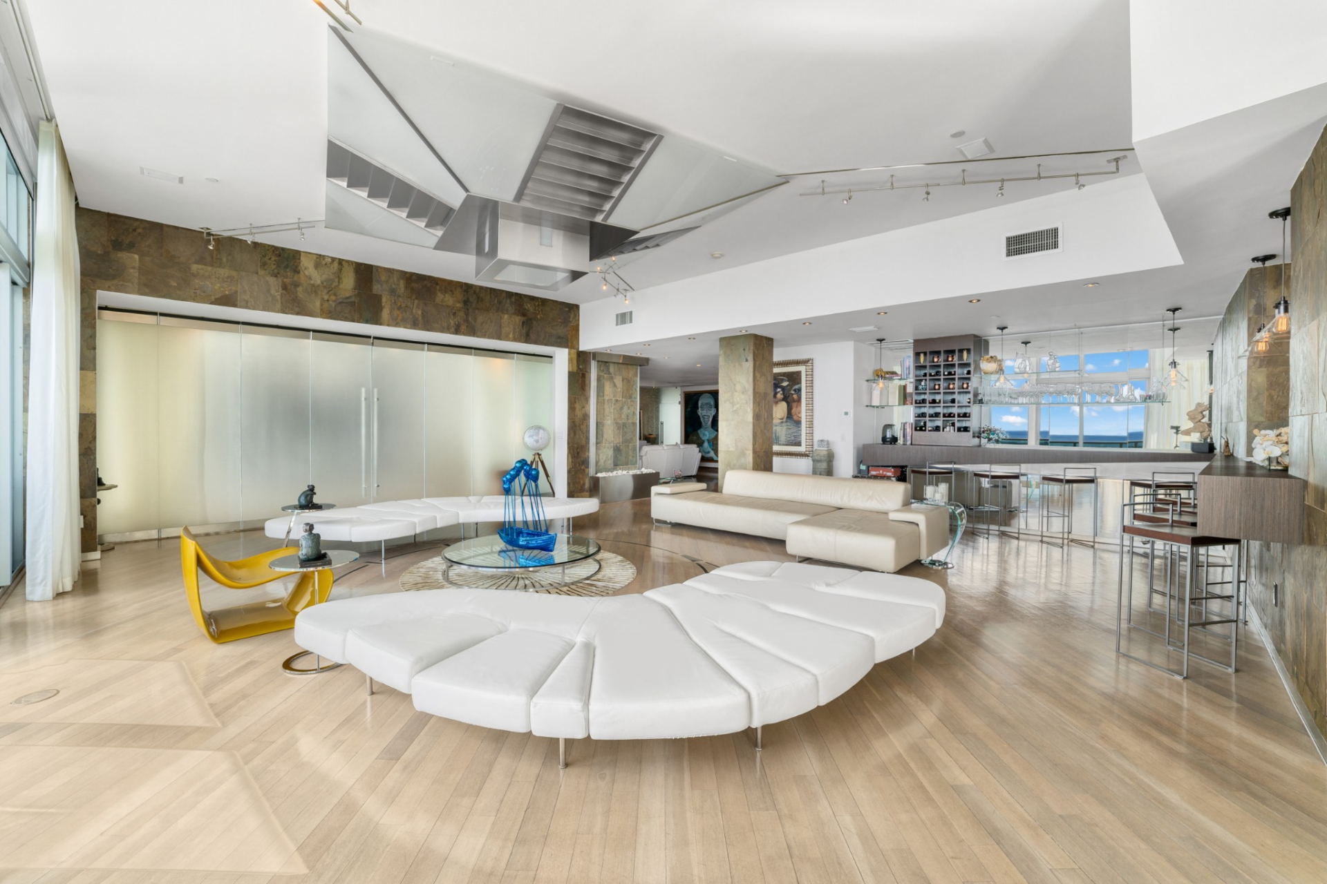 Wide-plank wood floors continue throughout, while oversized white seating pieces are arranged in sculptural formations, creating multiple conversation zones anchored by circular and glass coffee tables. 