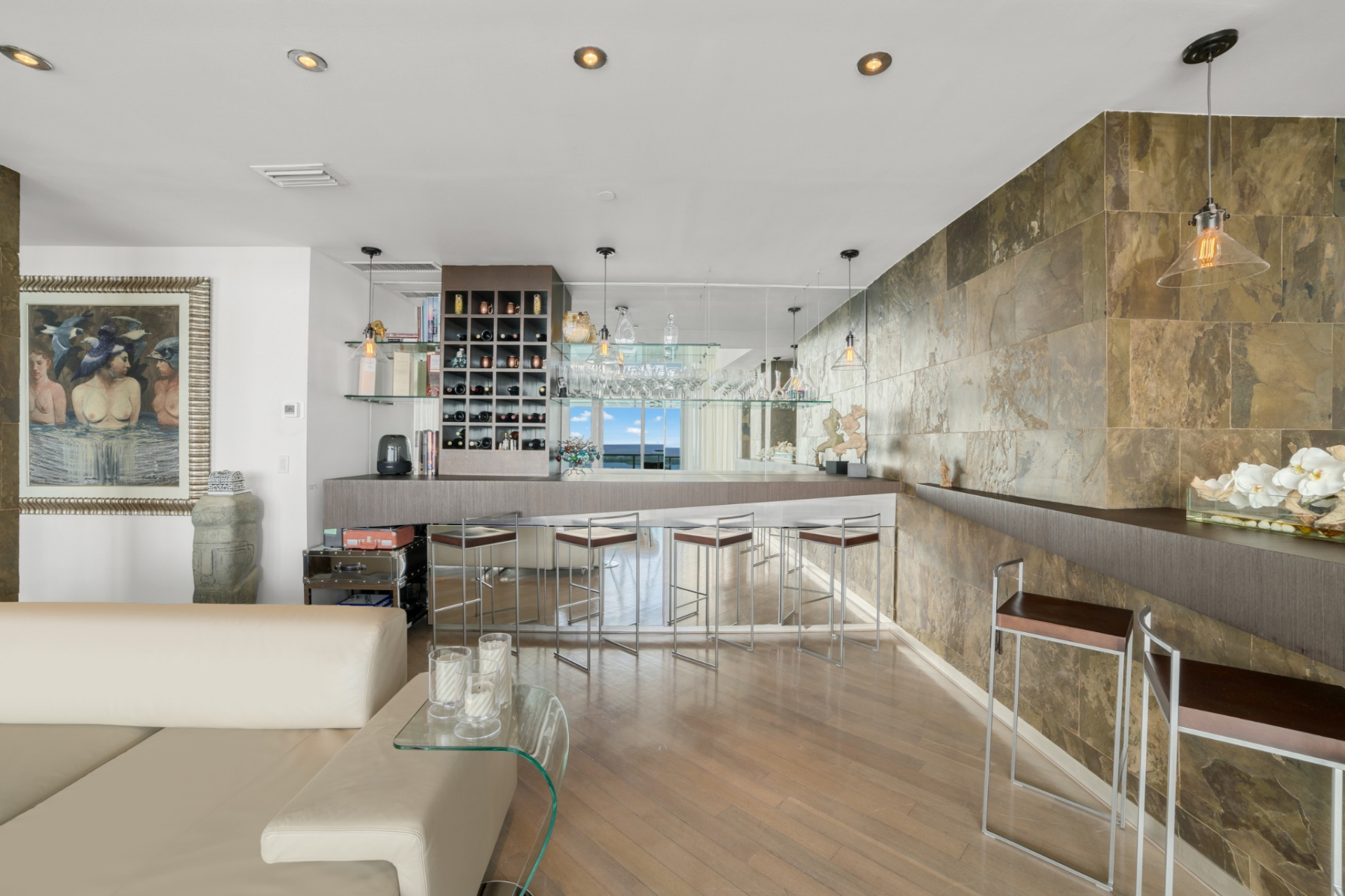 Along the back wall, a built-in bar features a long counter paired with metal-framed stools and glass shelving suspended from the ceiling, displaying stemware and spirits. 