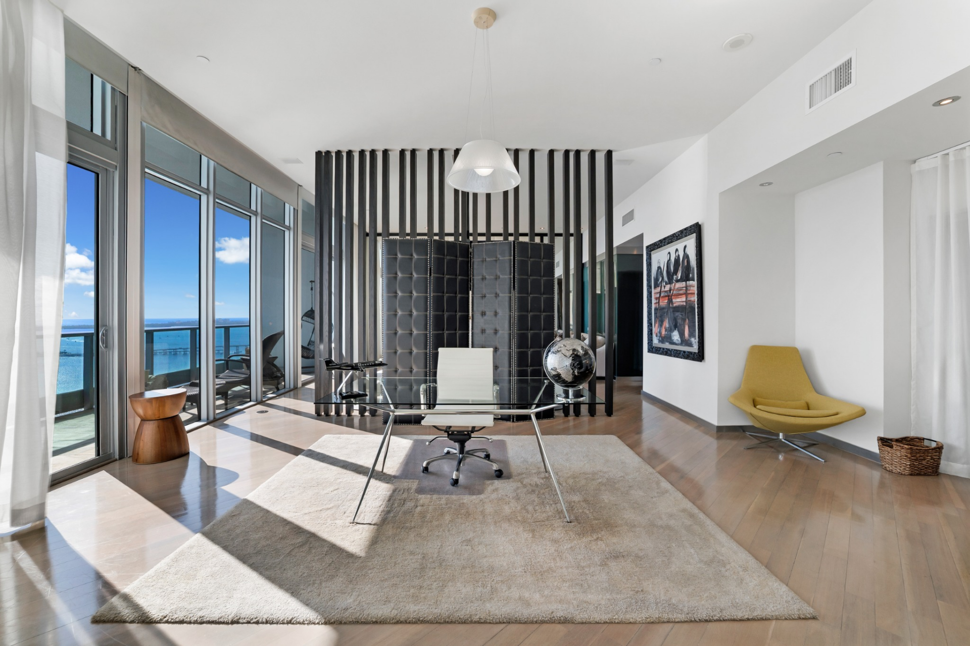 A sculptural black folding screen set against vertical slatted detailing separates the bedroom from the lounge, which currently features a glass-top desk with polished metal legs and a white chair.