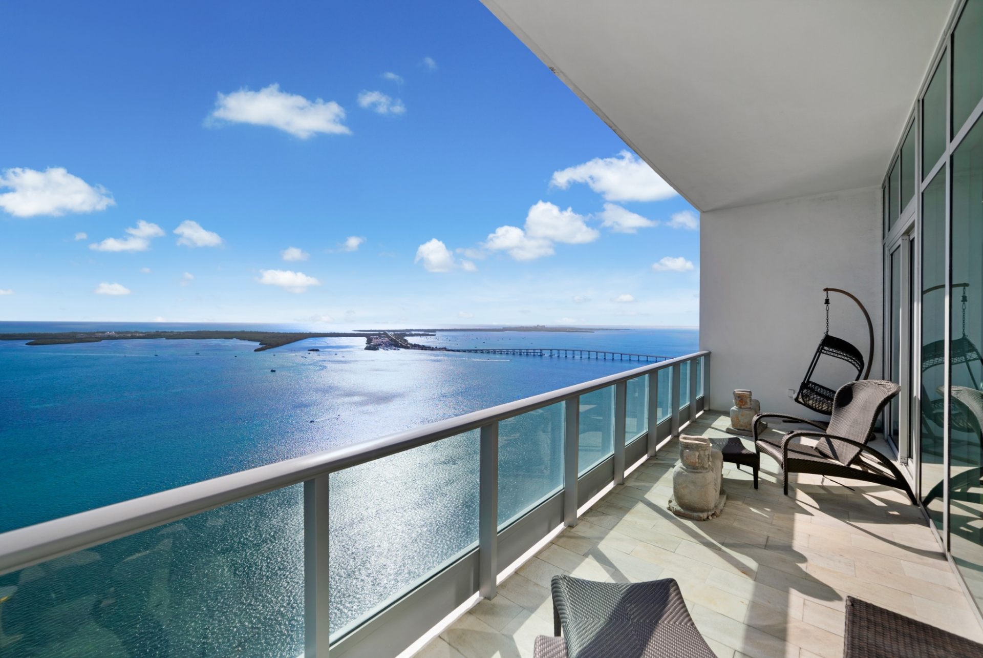 Woven lounge chairs and a suspended egg chair are arranged along the tiled balcony, positioned to take in the open skyline and the reflective surface of the water below.