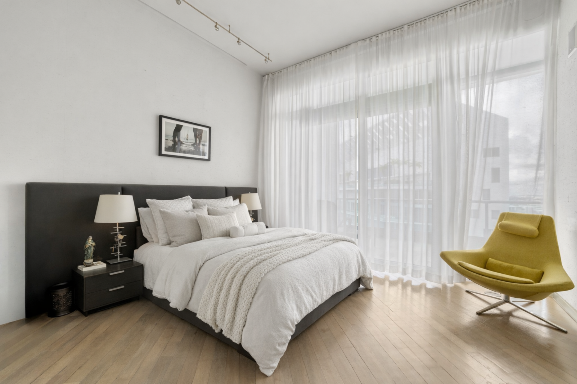 The bedroom features a dark upholstered headboard that contrasts with crisp white bedding layered with textured throws and structured pillows. 
