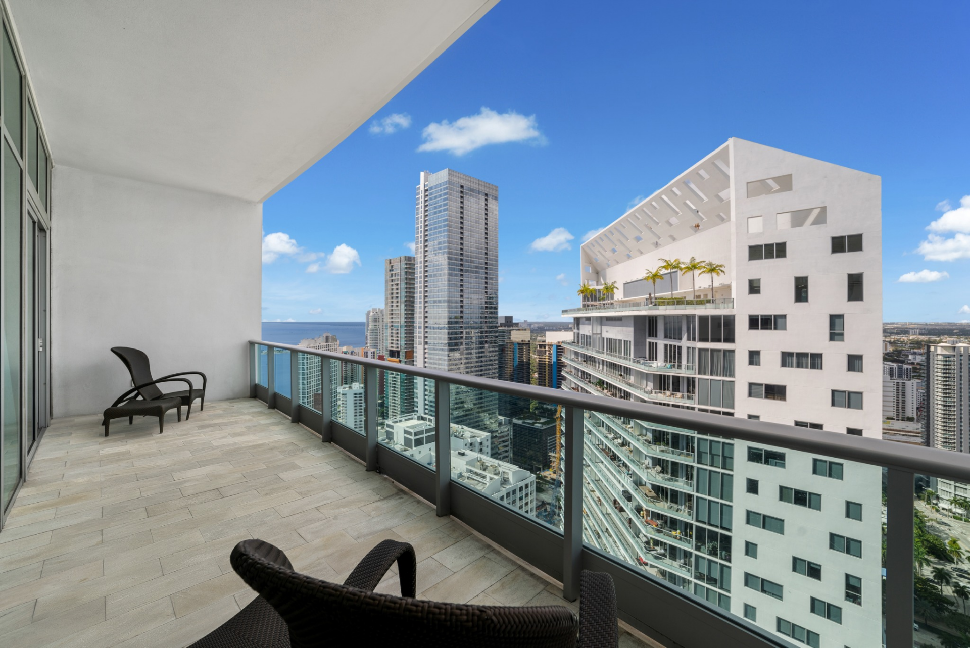 In an outdoor balcony, glass railings frame views of neighboring high-rises and a stretch of blue water beyond, with woven outdoor chairs positioned along the tiled balcony.
