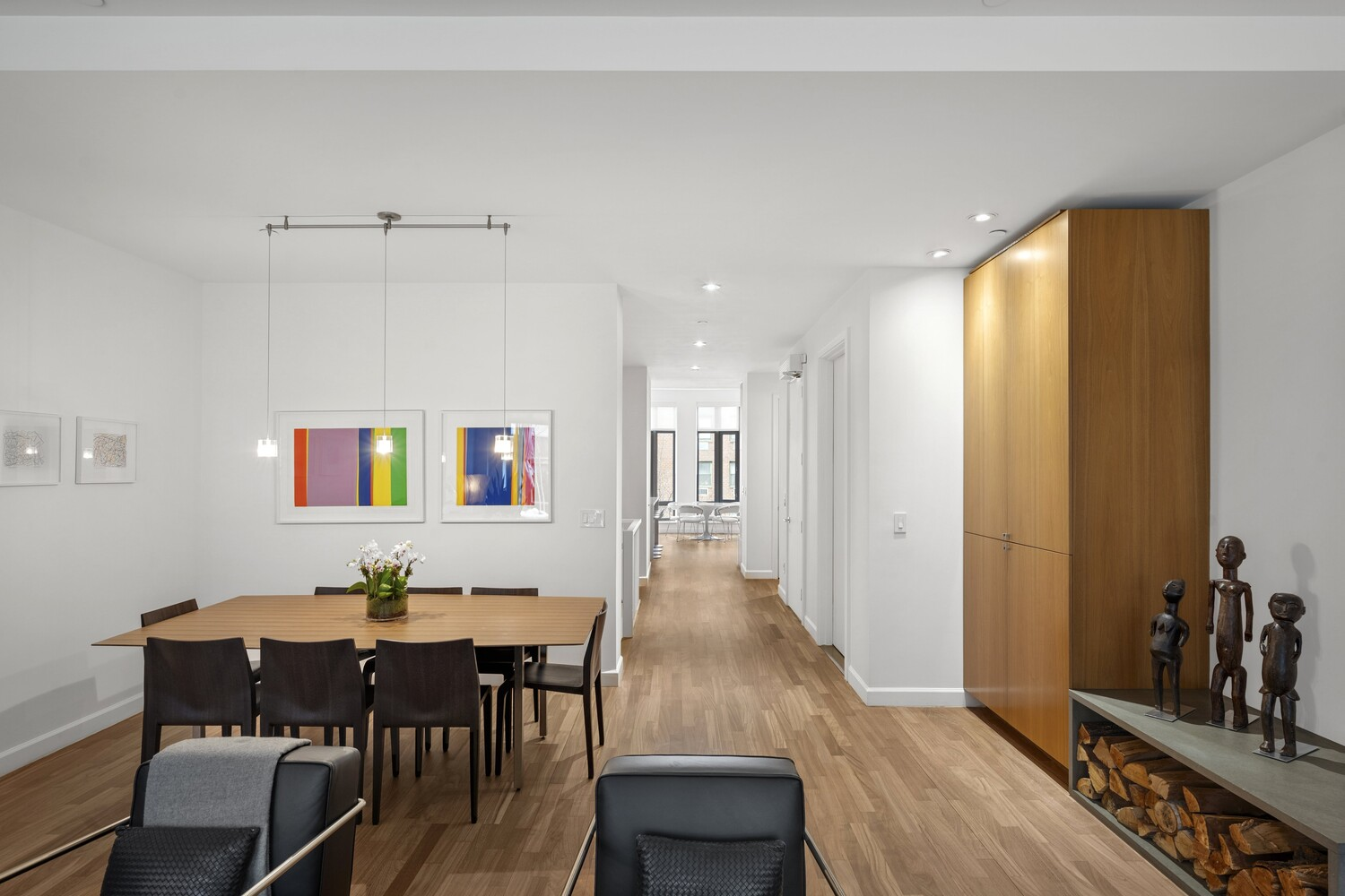 A long rectangular dining table sits in a left corner, positioned beneath a pair of slim, suspended pendant lights that drop from the ceiling. 