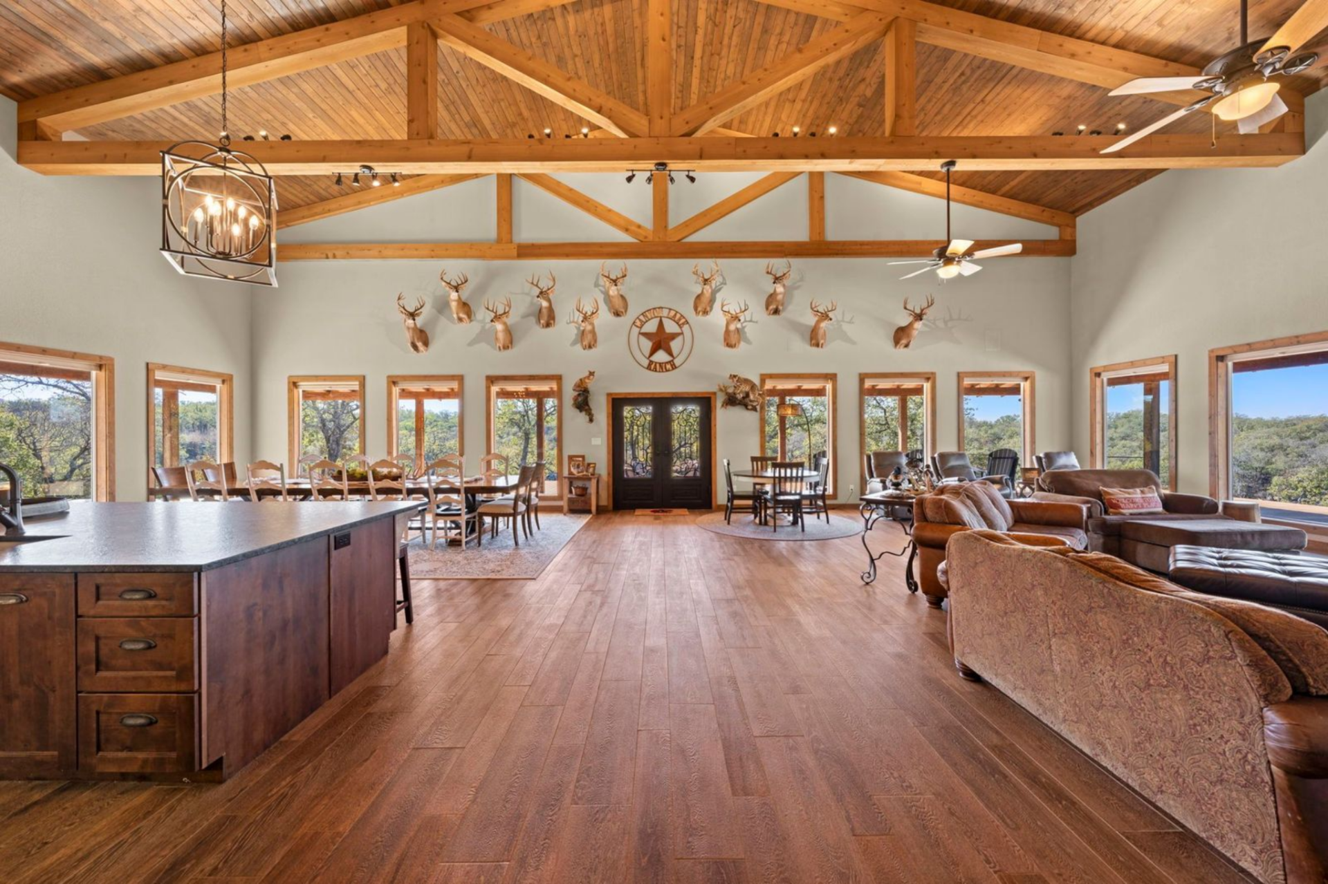 The space unfolds in one continuous line of sight, from the kitchen island in the foreground to the dining area and into the living room, all framed by warm wood floors and exposed timber trusses overhead.