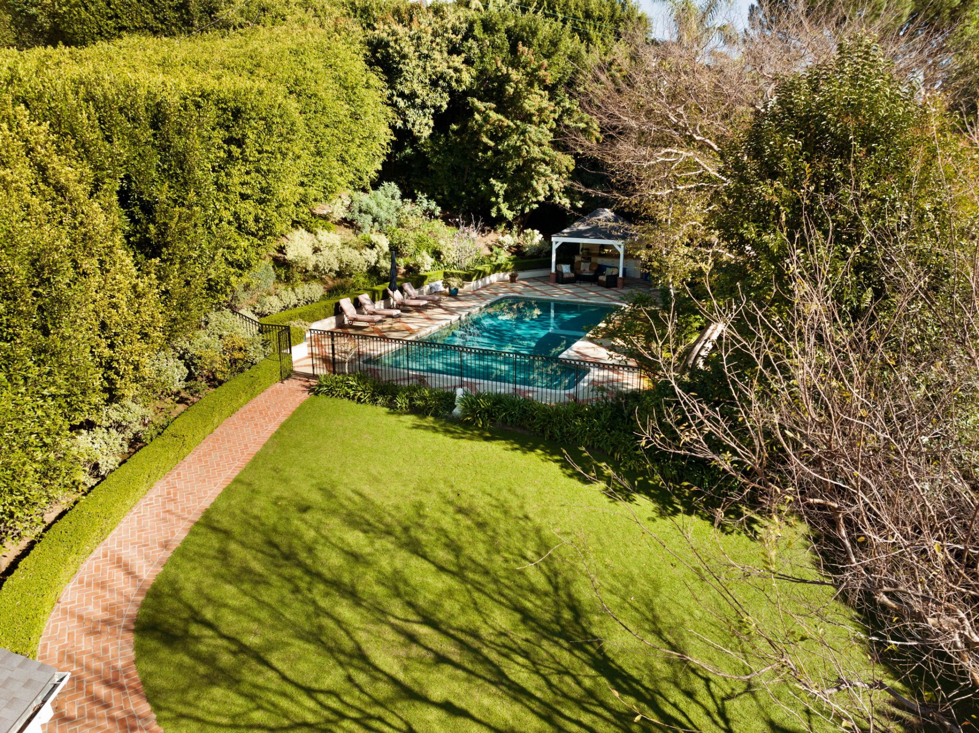A brick pathway curves gently through the grass, guiding you toward the far end of the yard where a gated swimmer’s pool and cabana complete the property.