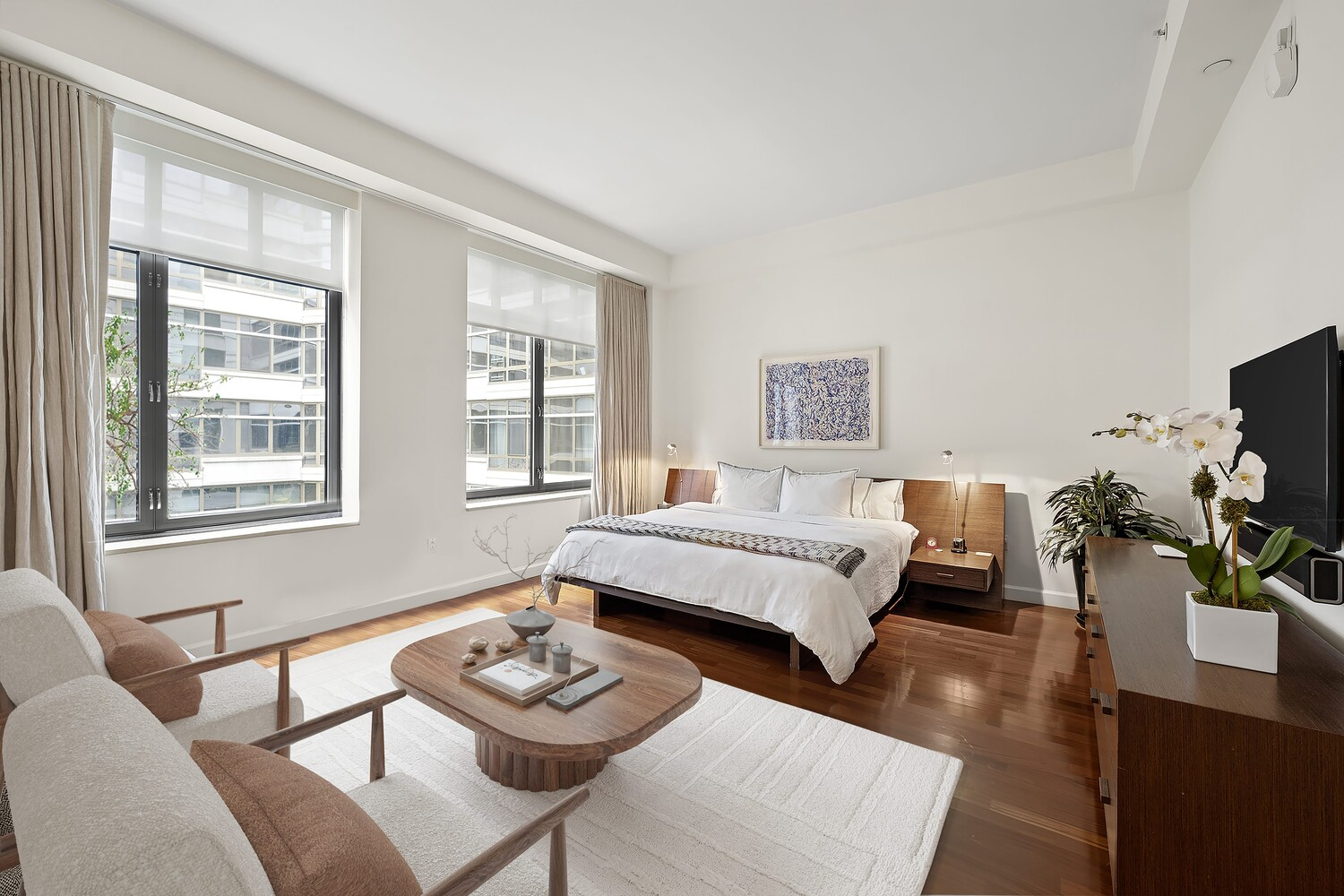 Large windows run along the exterior wall, while inside the room is a bed, a sitting area, and a TV mounted above a dresser.