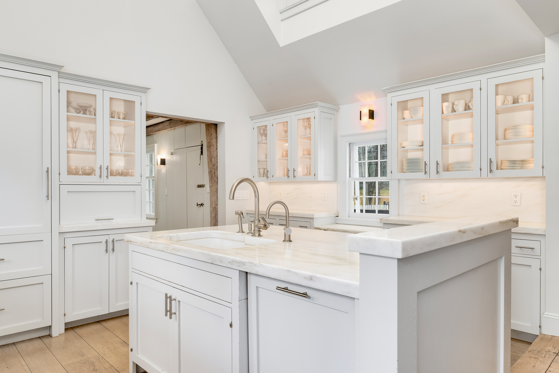Glass-front upper cabinets display neatly arranged glassware and dishes and a professional-grade range sits below a tailored hood surround framed by windows.