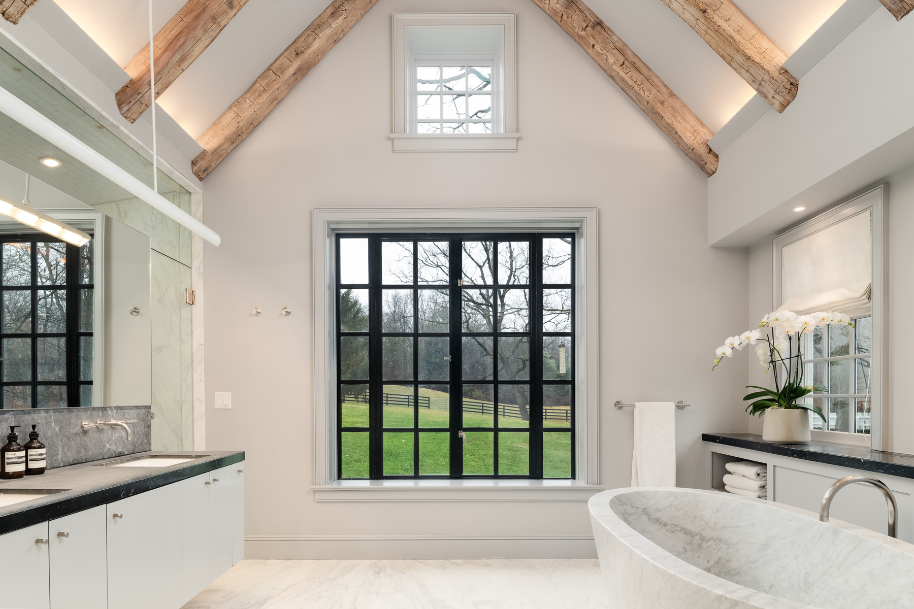 The en-suite bathroom continues the suite’s refined aesthetic with a large window in the center, positioned to capture sweeping views of the property’s rolling lawn. 