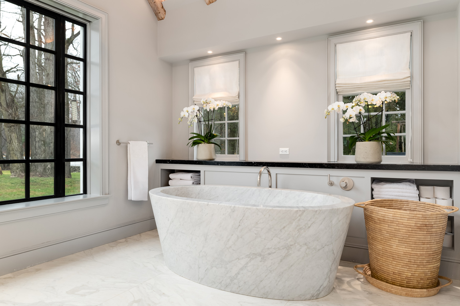 To one side of the window, a sculptural freestanding marble soaking tub sits in front of built-in cabinetry meant to store towels and toilet paper.