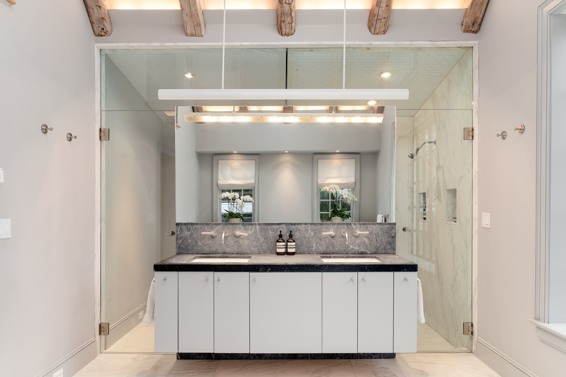 On the other side, a long dual vanity with stone countertops and integrated sinks is framed by a full glass enclosure that houses the shower area.