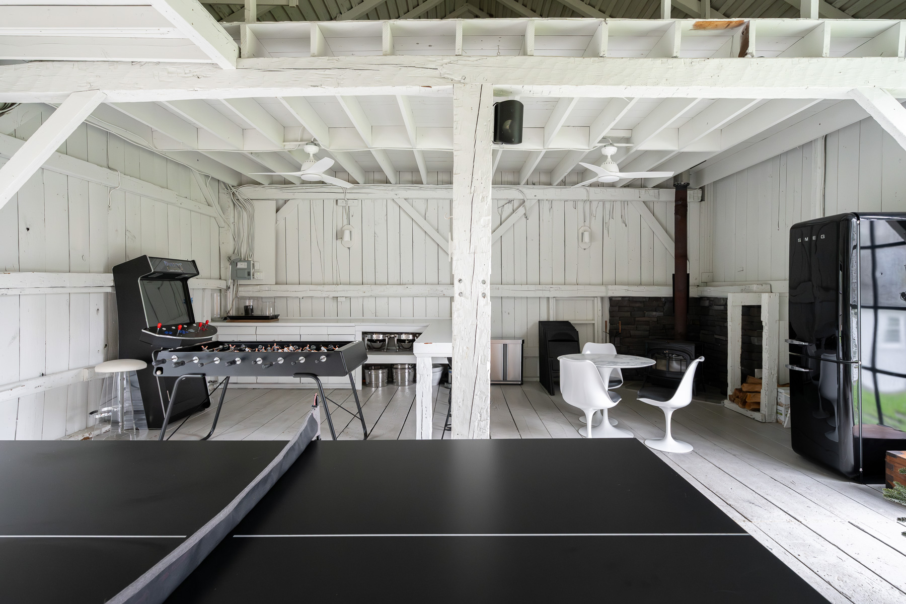 Inside, one side of the barn features a ping-pong table and arcade setup, turning it into a casual game lounge, while the other features a seating area anchored by a wood-burning fireplace and a small kitchenette.
