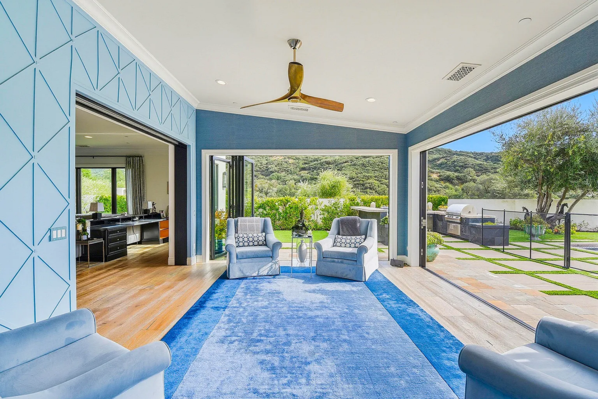 In the space, a pair of lounge chairs rest on a saturated blue rug beneath a modern ceiling fan, creating a shaded sitting area that feels fully connected to the landscape beyond.