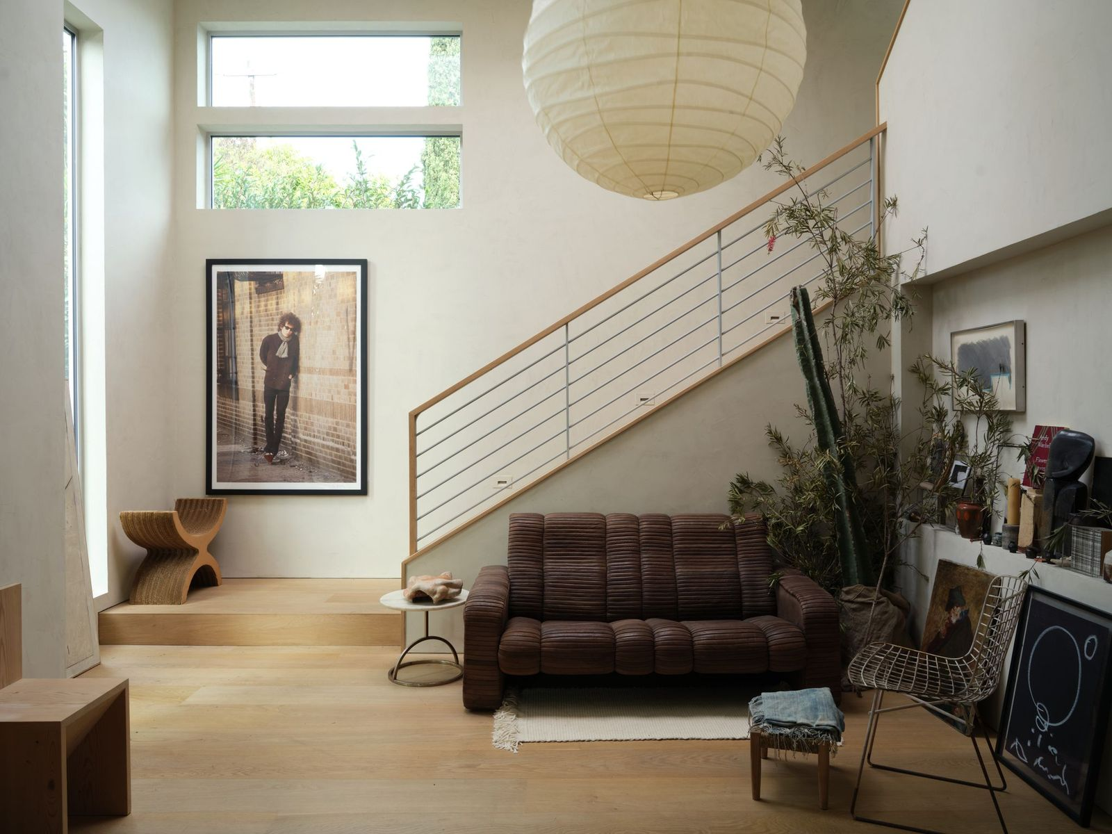 Just to the right of the entrance, a relaxed sitting area unfolds beneath the soaring ceiling. A low, ribbed brown sofa is paired with a simple round side table, art, plants, and collected objects that keep the look understated but intentional.
